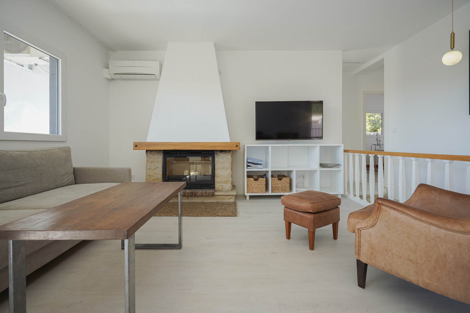 Salon lumineux avec cheminée en pierre, climatisation réversible, mobilier en cuir et escalier avec balustrade blanche et bois.