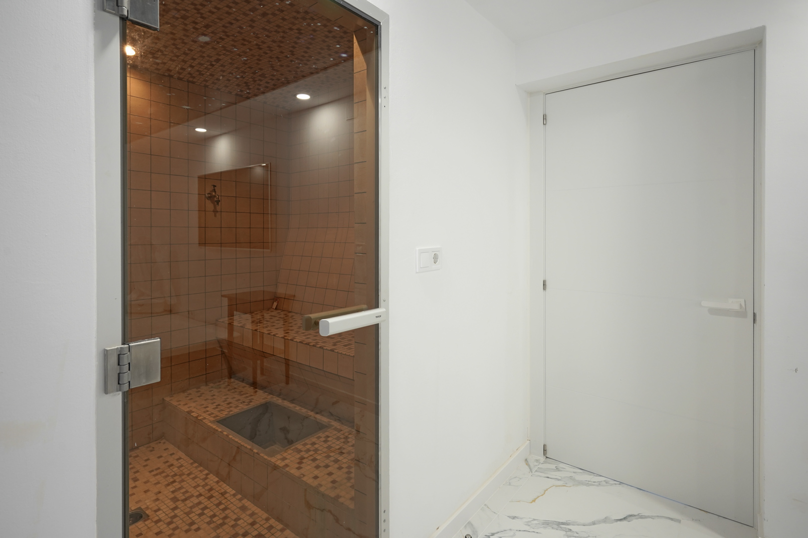 Sauna privé avec revêtement en mosaïque céramique, éclairage encastré et porte vitrée, situé à côté d'une pièce au sol en marbre blanc.