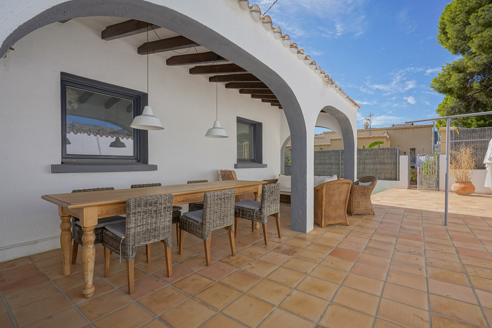Terrasse couverte avec arches blanches, poutres apparentes et sol en terre cuite. Elle dispose d'un coin repas avec table en bois et chaises en osier.