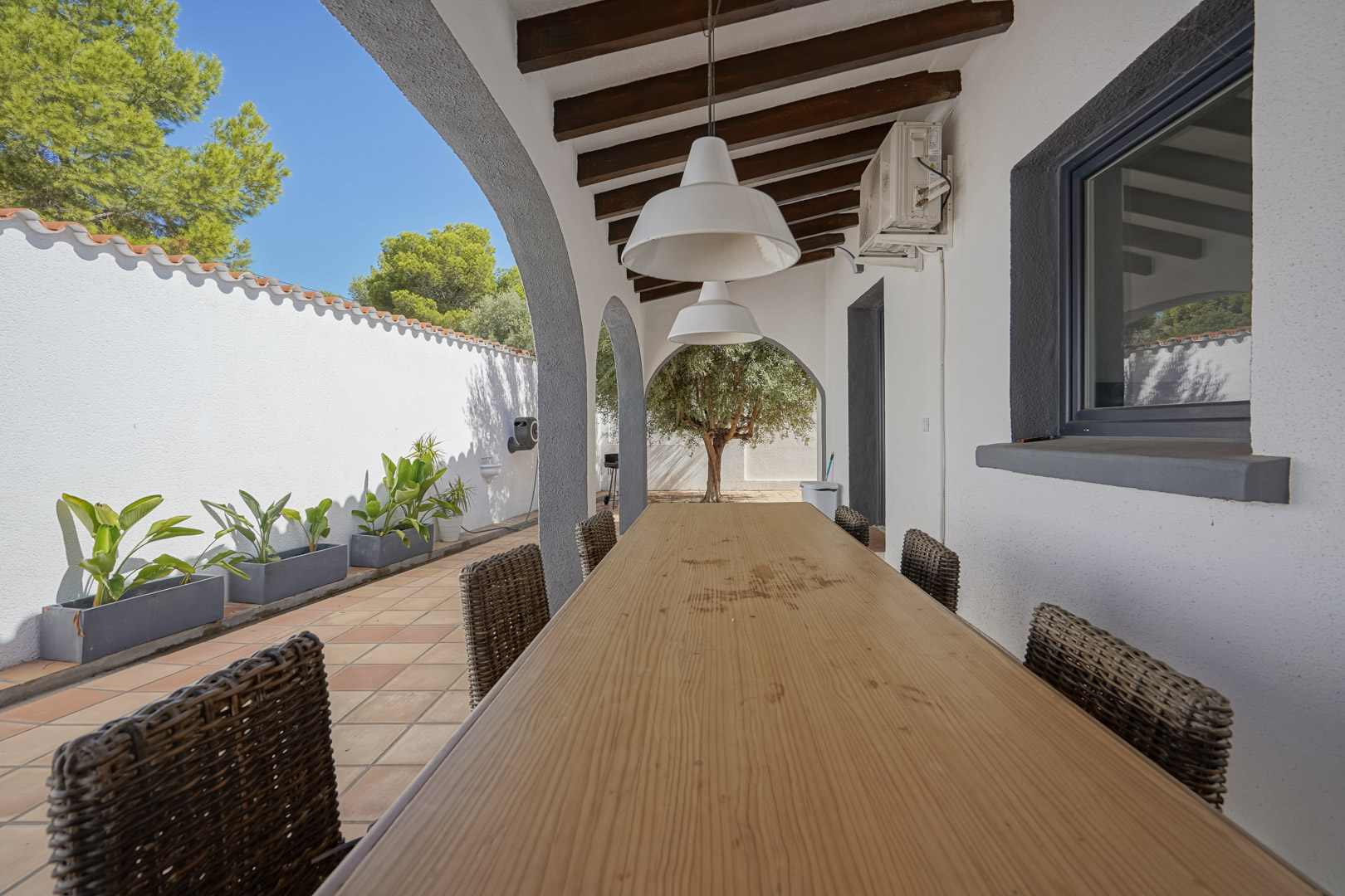 Terrasse couverte avec poutres apparentes, grande table à manger en bois et chaises en osier. L'espace s'ouvre sur un jardin méditerranéen par des arches élégantes.