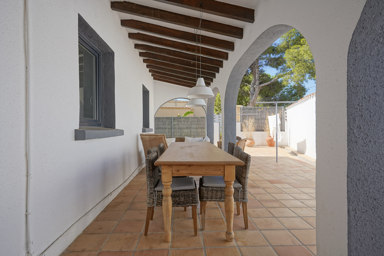 Terrasse couverte avec poutres en bois apparentes, arches méditerranéennes et coin repas extérieur avec table en bois et chaises en osier sur sol en terre cuite.