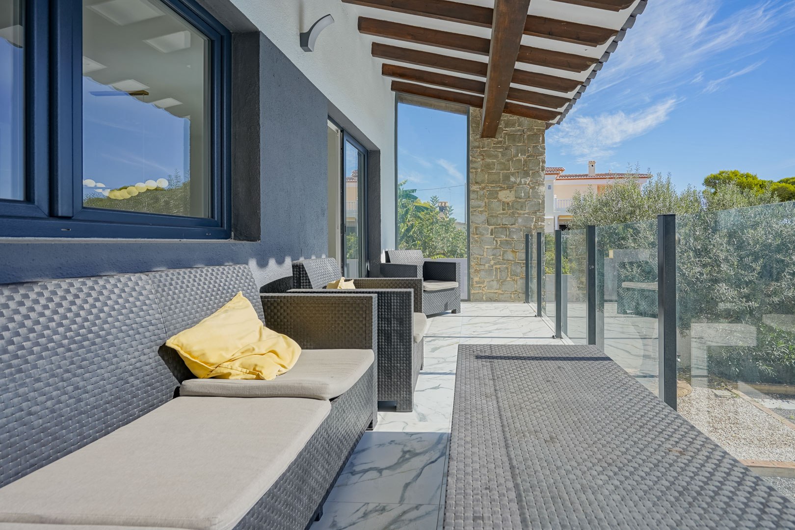 Terrasse couverte avec poutres en bois apparentes, mobilier en rotin synthétique et sol en marbre. Un garde-corps en verre offre une vue sur le jardin méditerranéen.