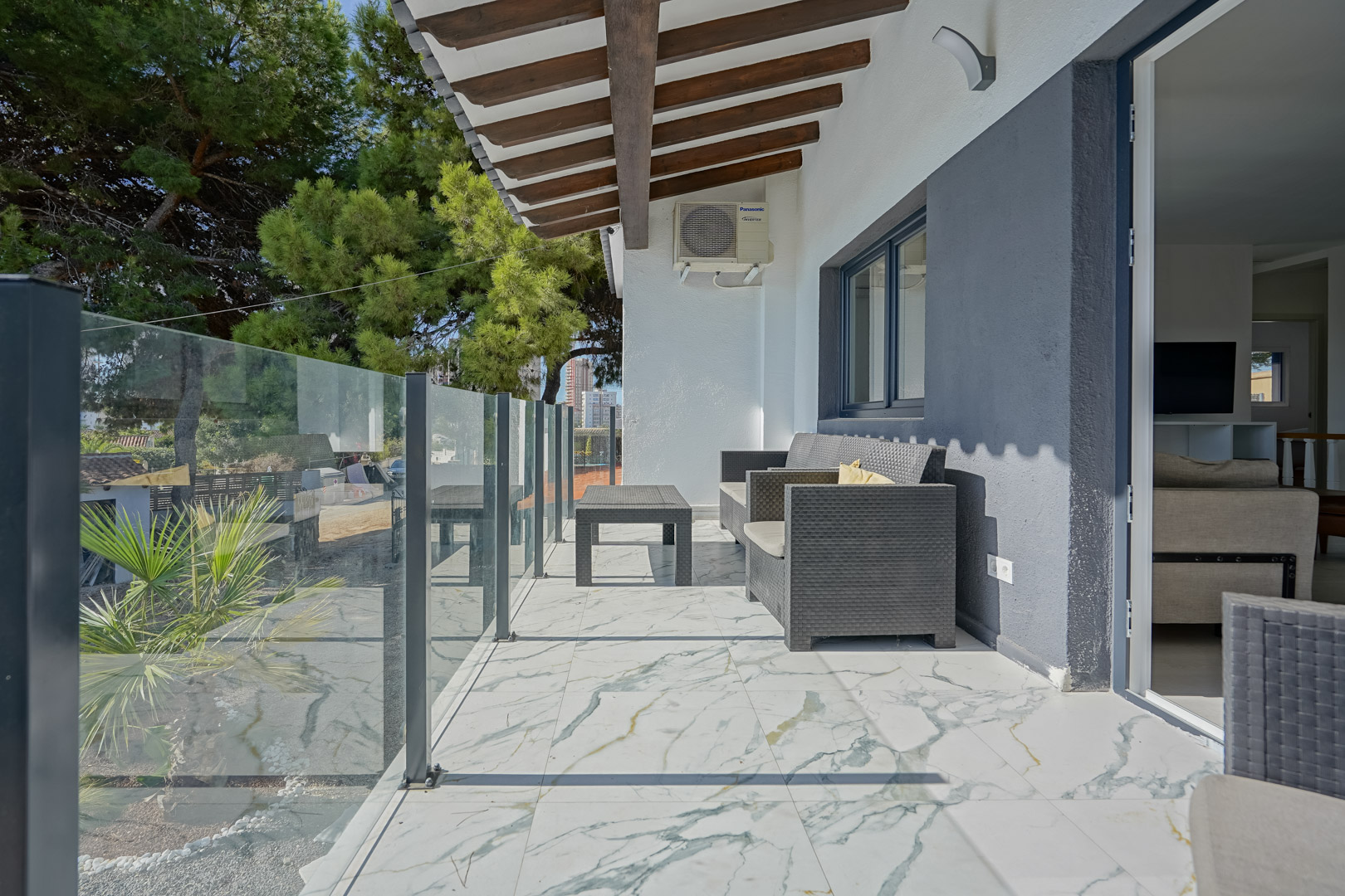 Terrasse couverte avec sol en marbre veiné, garde-corps en verre et plafond à poutres en bois. Meublée de rotin gris, elle offre une vue sur le jardin arboré.