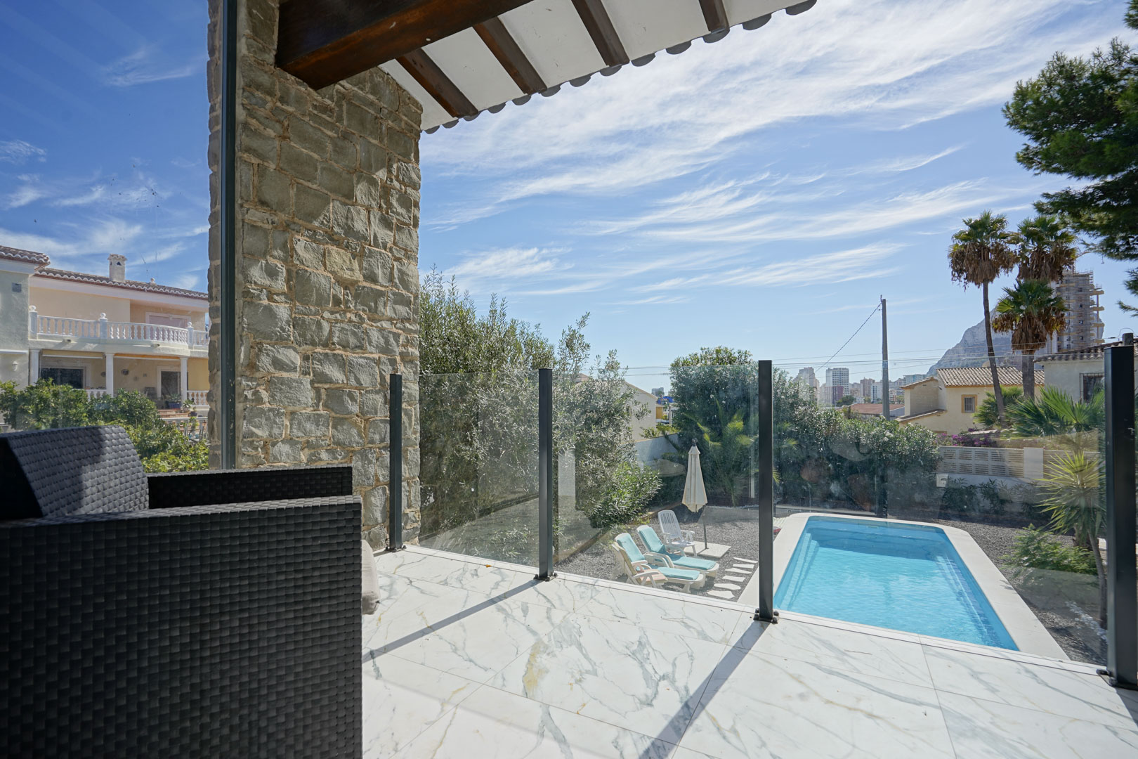 Terrasse supérieure avec carrelage effet marbre, garde-corps en verre et vue sur la piscine privée, le jardin et le rocher d'Ifach au loin.