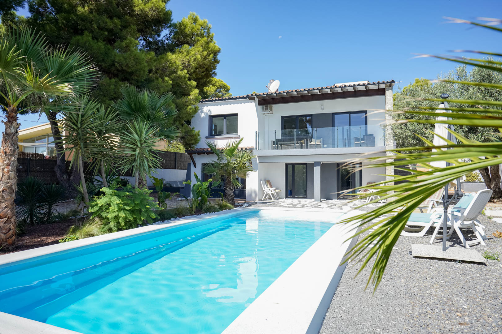 Vue extérieure de la villa avec piscine rectangulaire, jardin méditerranéen arboré et façade blanche à deux niveaux avec terrasses ensoleillées.