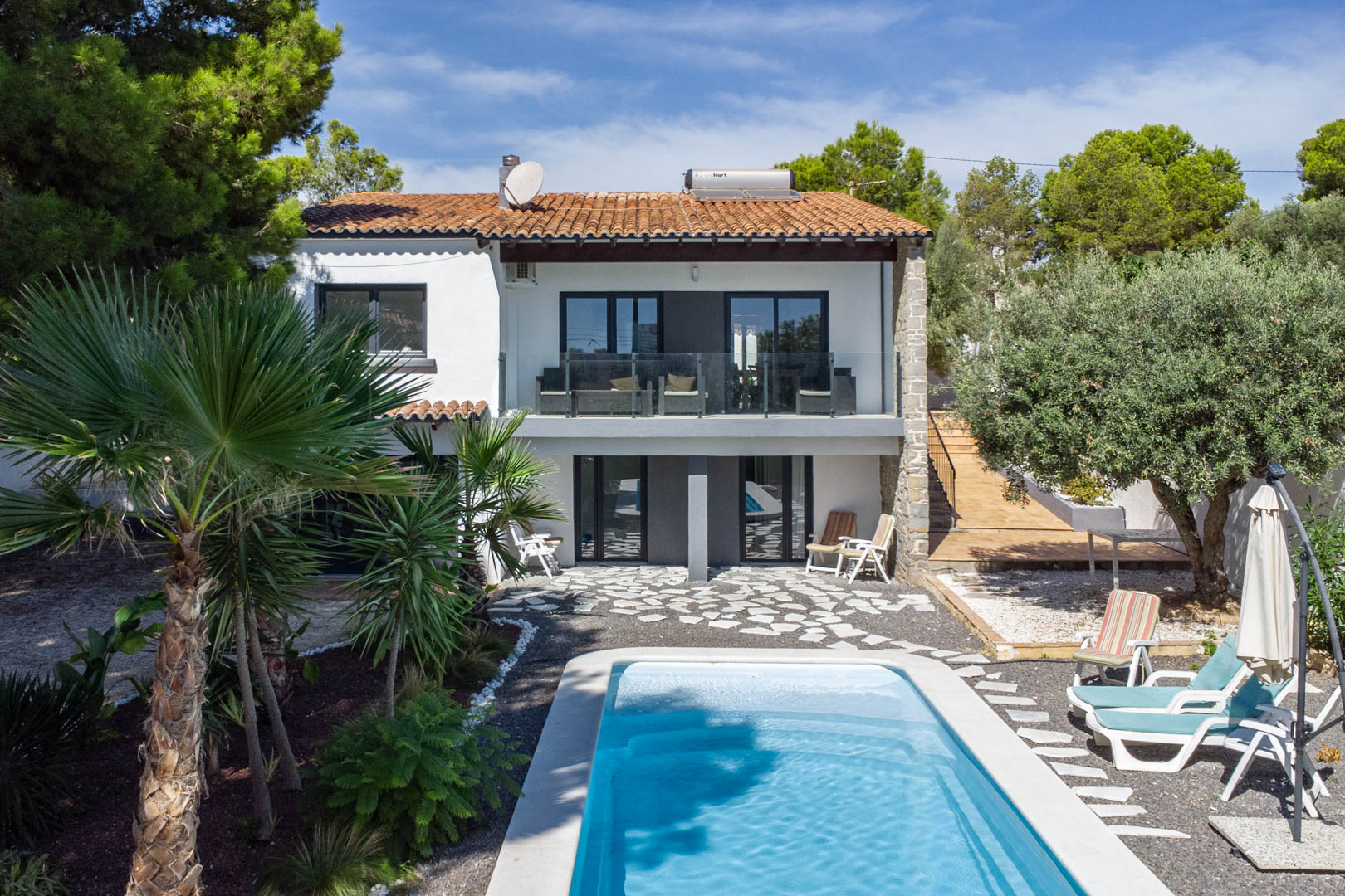 Vue extérieure de la villa avec piscine rectangulaire, jardin méditerranéen et façade blanche. Comprend des terrasses avec garde-corps en verre et un chauffe-eau solaire.