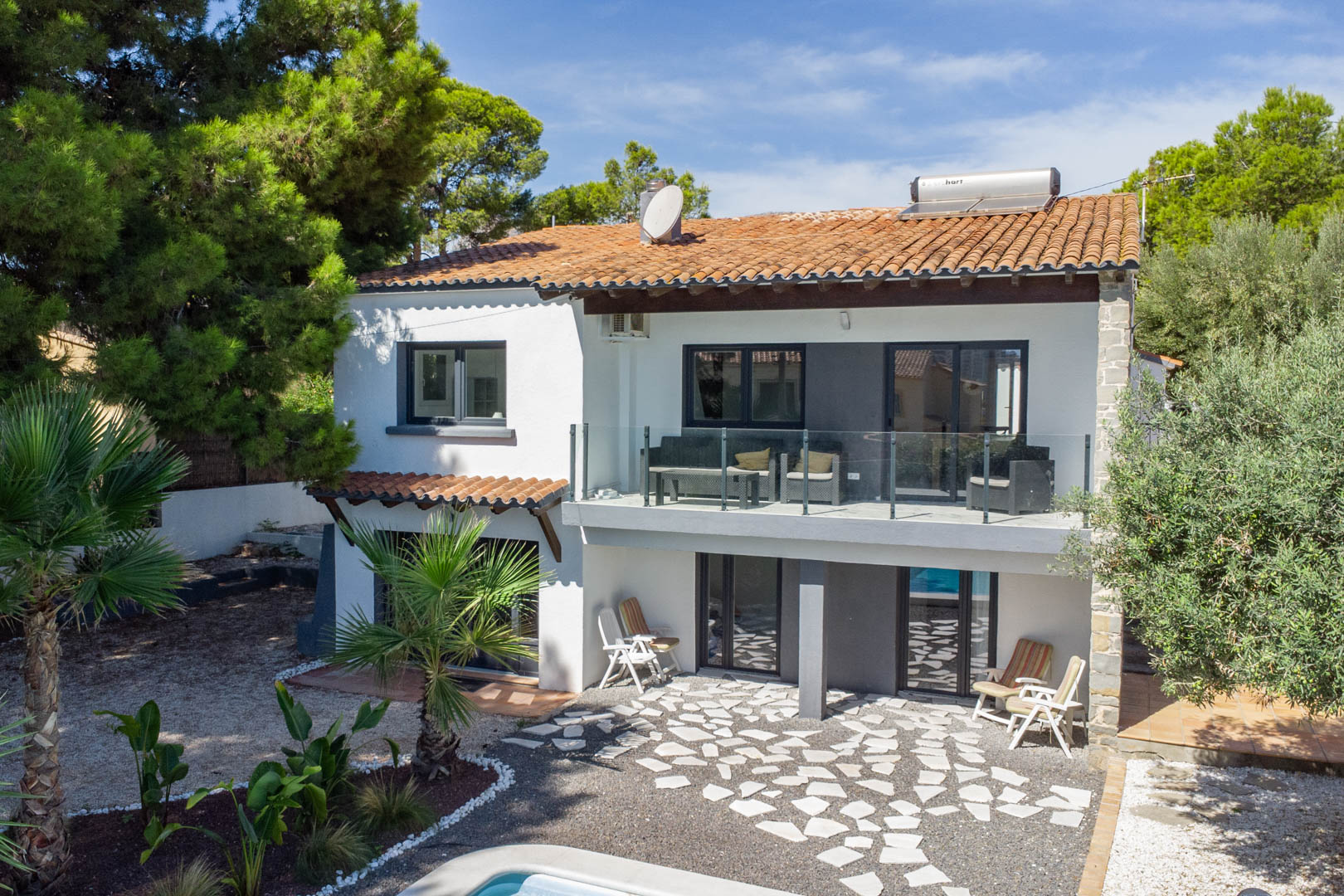 Vue extérieure de la villa sur deux niveaux avec terrasse vitrée, chauffe-eau solaire sur le toit et patio inférieur pavé de pierres naturelles.
