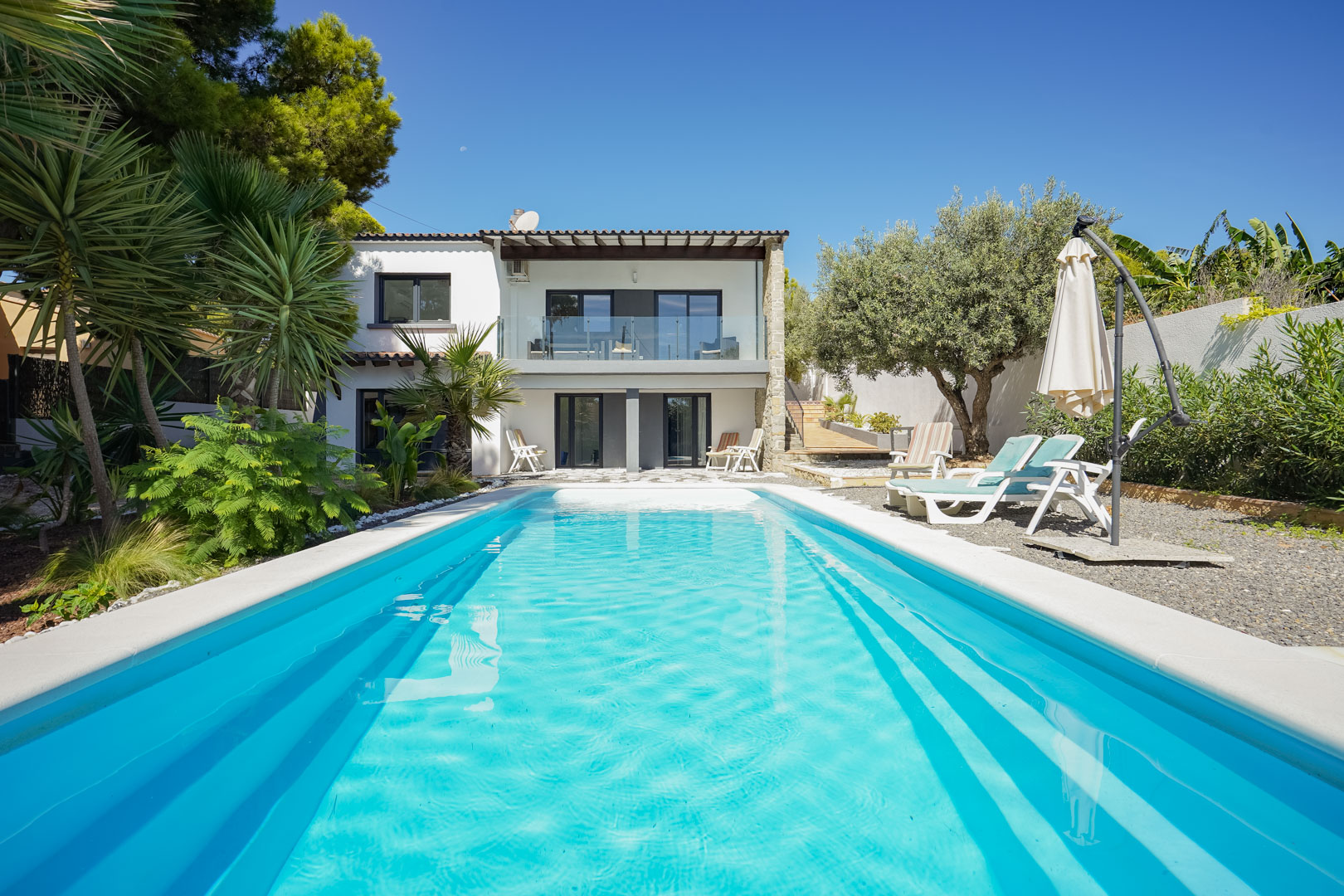 Vue sur la piscine rectangulaire et le jardin méditerranéen avec palmiers, illustrant la façade blanche de la villa et sa terrasse à balustrade en verre.