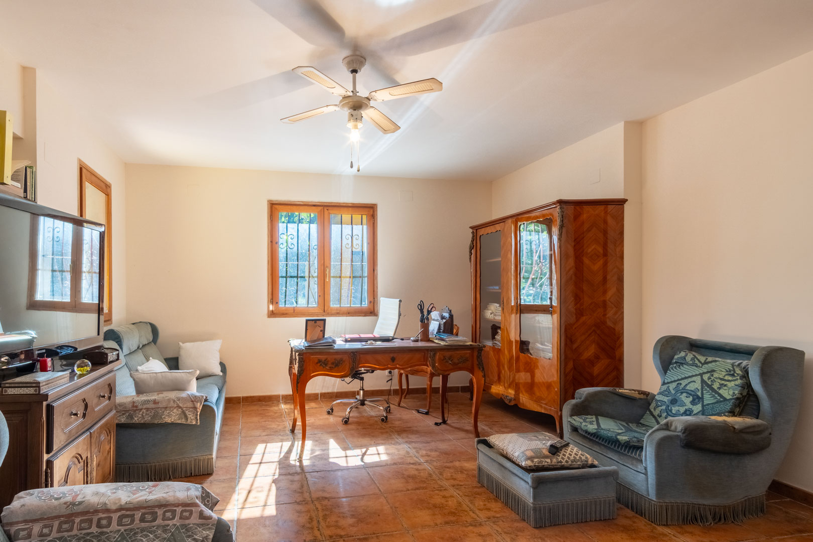 Bureau lumineux avec sol en terre cuite, bureau en bois classique et armoire assortie. La pièce dispose d'un ventilateur de plafond et de fauteuils rembourrés.