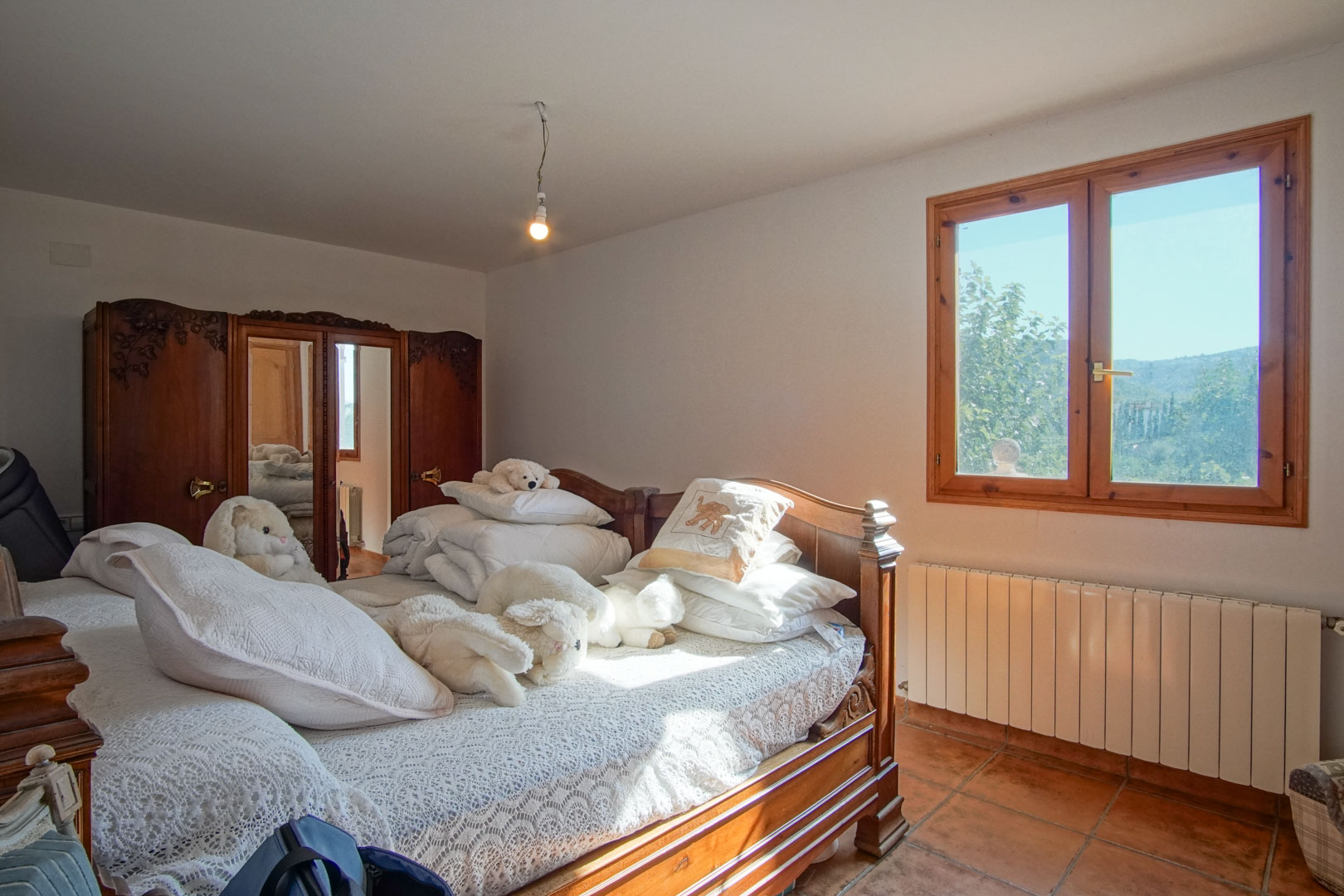 Chambre avec mobilier en bois sculpté, armoire à miroirs, sol en terre cuite et fenêtre offrant une vue sur la nature, équipée d'un radiateur.