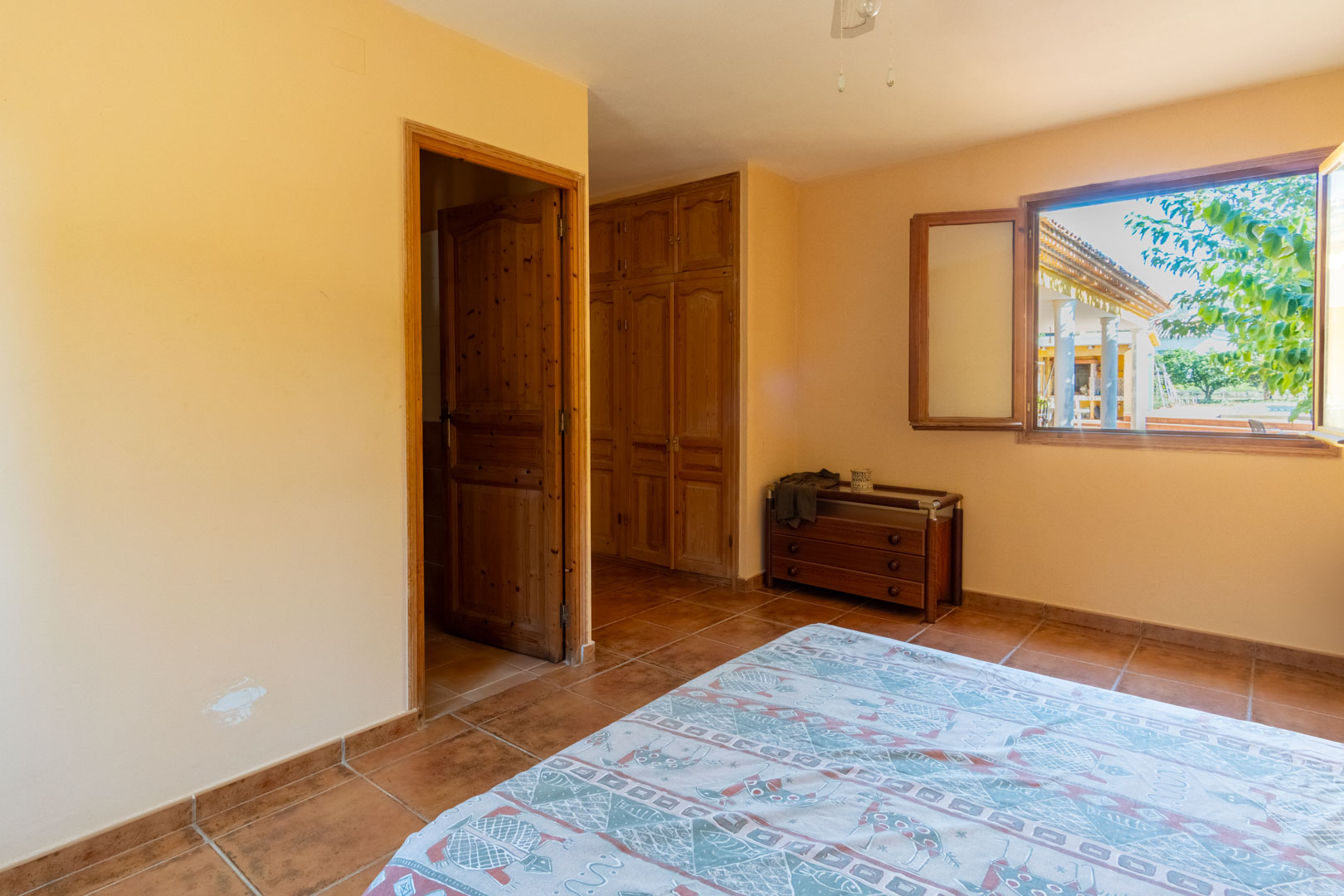 Chambre avec sol en terre cuite, armoires encastrées en bois massif et fenêtre ouvrant sur le porche extérieur et la végétation.