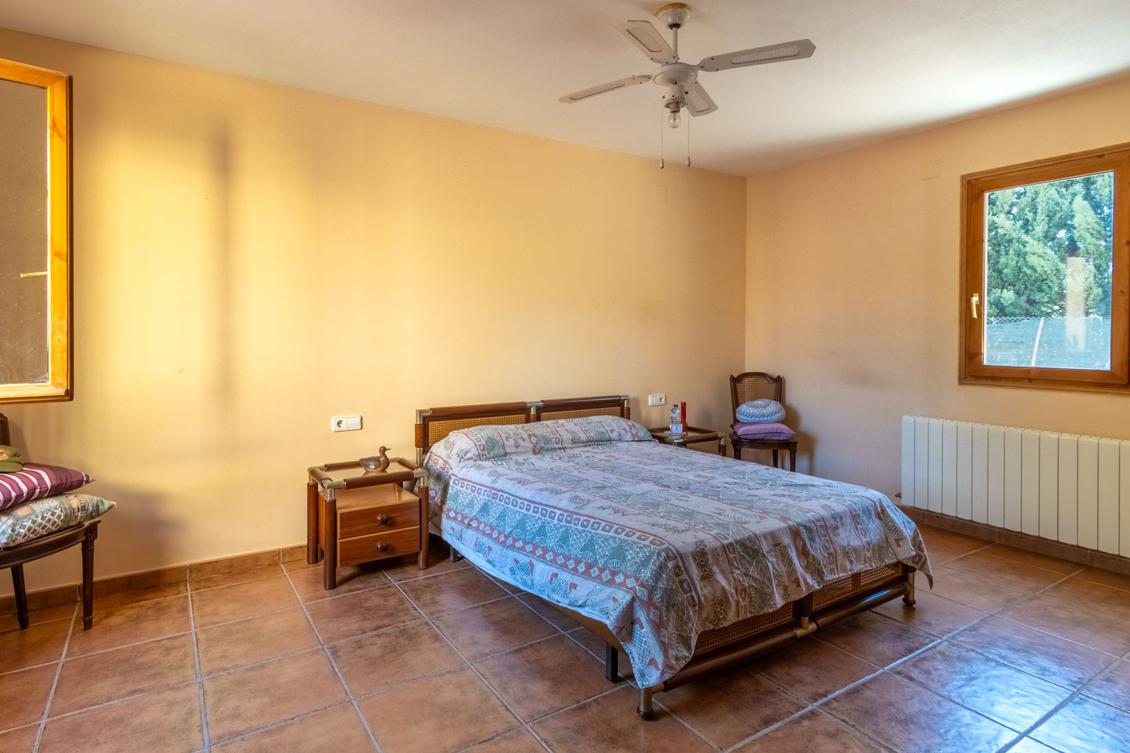 Chambre avec sol en terre cuite, murs aux tons chauds et ventilateur de plafond. Elle dispose de fenêtres à cadre en bois, d'un radiateur et de tables de chevet en bois assorties.