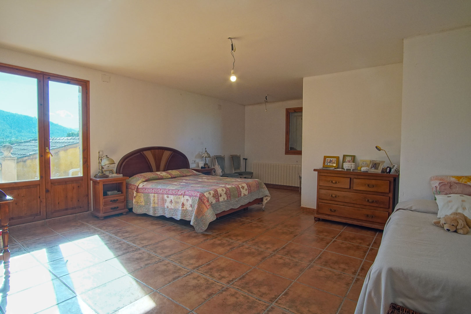 Chambre spacieuse avec sol en terre cuite, mobilier en bois classique et porte vitrée offrant une vue dégagée sur les montagnes environnantes.