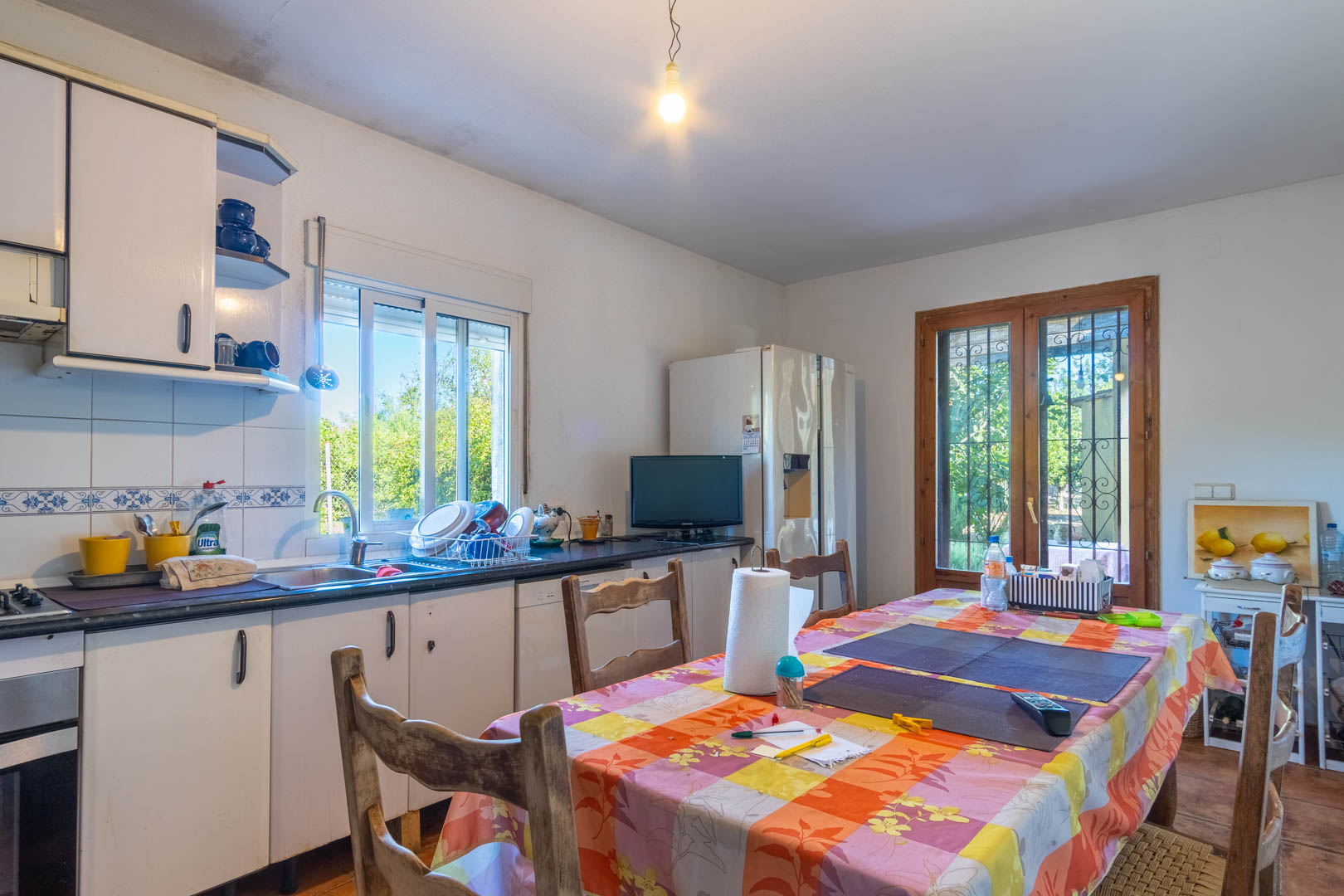 Cuisine fonctionnelle avec placards blancs, plan de travail sombre et coin repas. Elle dispose d'un accès direct à l'extérieur et de vues sur le jardin via des menuiseries en bois.