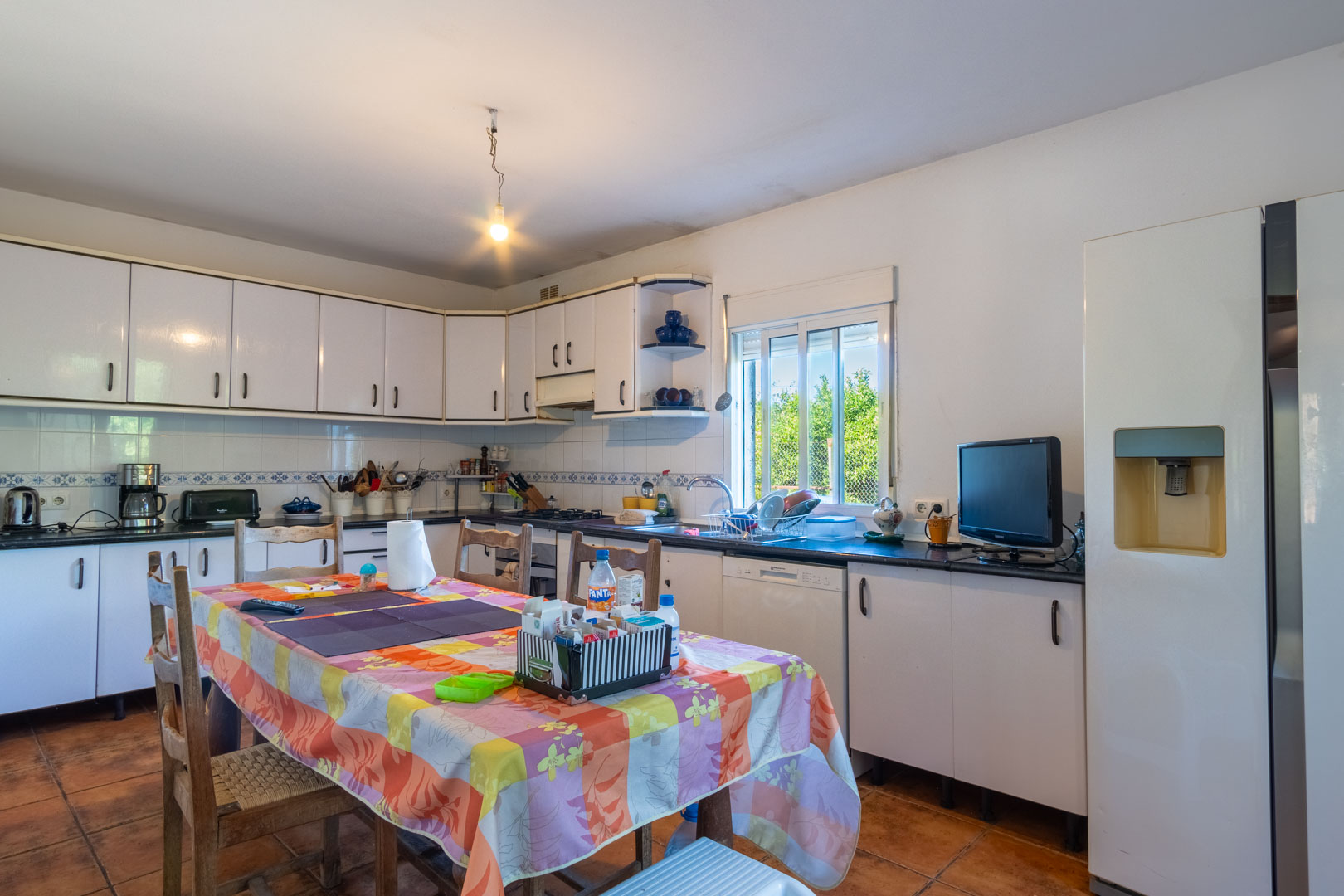 Cuisine indépendante avec meubles blancs, plan de travail sombre et coin repas. Elle dispose d'un sol en céramique et d'une fenêtre offrant une vue sur le jardin.