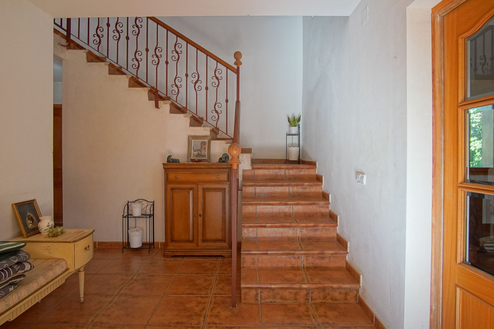 Escalier intérieur avec marches en terre cuite et rampe en fer forgé travaillé. Le hall d'entrée présente des sols rustiques et du mobilier en bois de style classique.