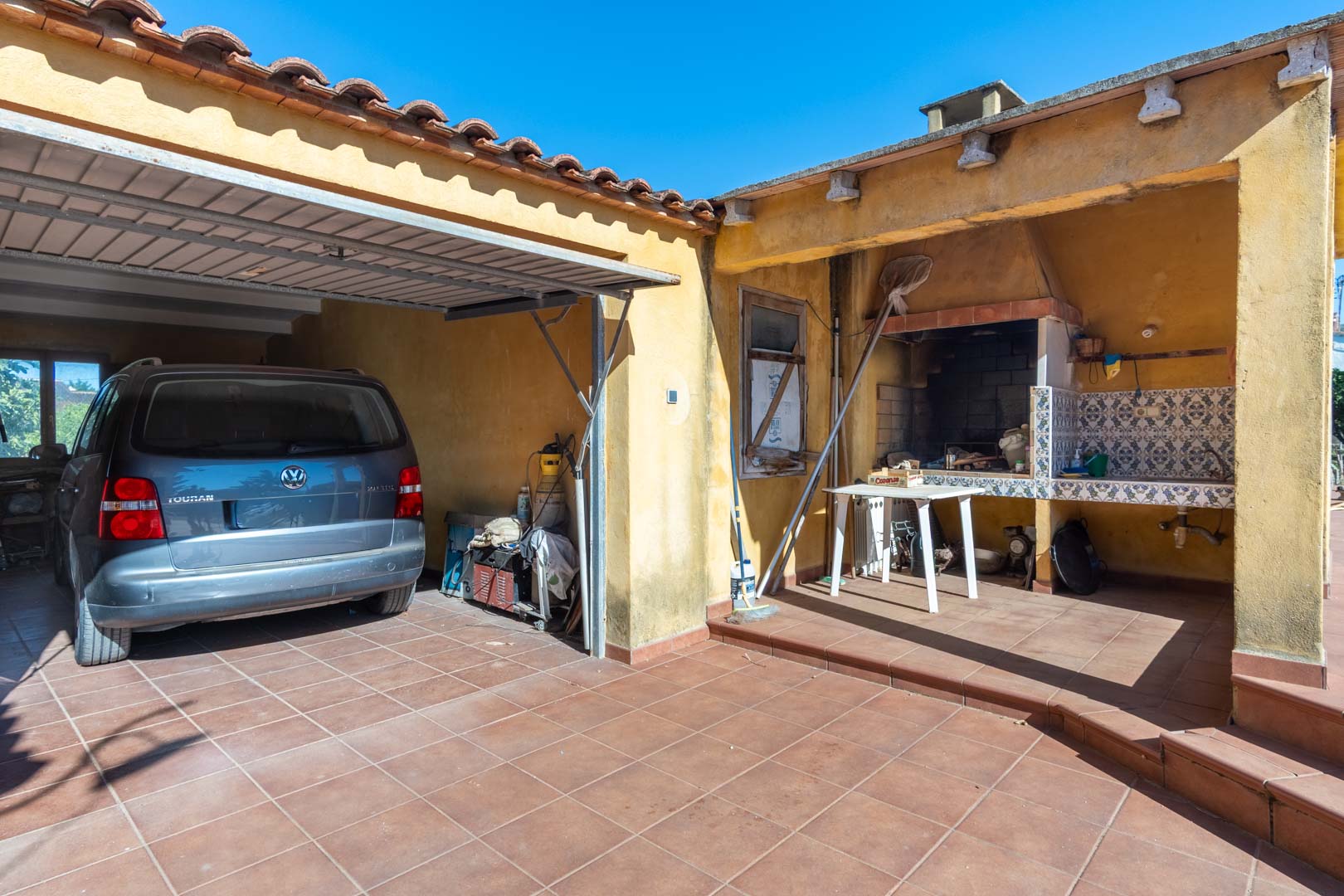 Espace extérieur fonctionnel comprenant un parking couvert et un coin barbecue traditionnel avec plan de travail carrelé et évier.