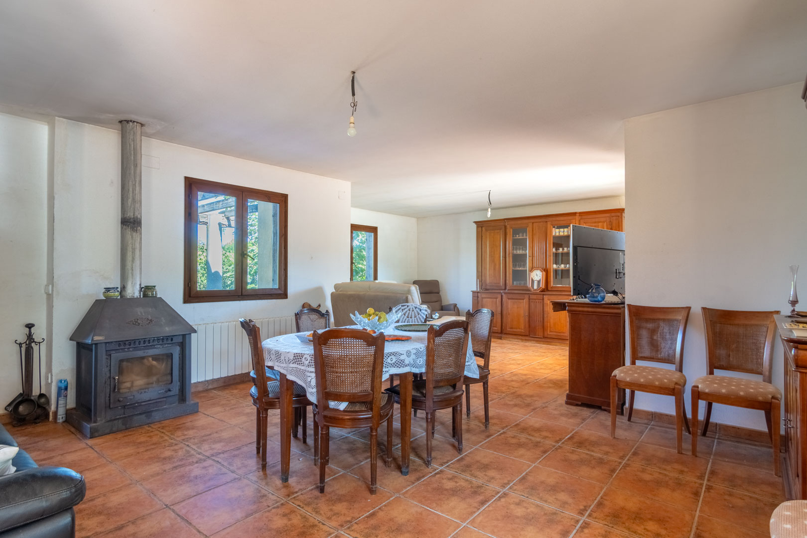 Espace salon-salle à manger avec sol en grès rustique, poêle à bois métallique et mobilier classique en bois incluant un grand buffet vitré.