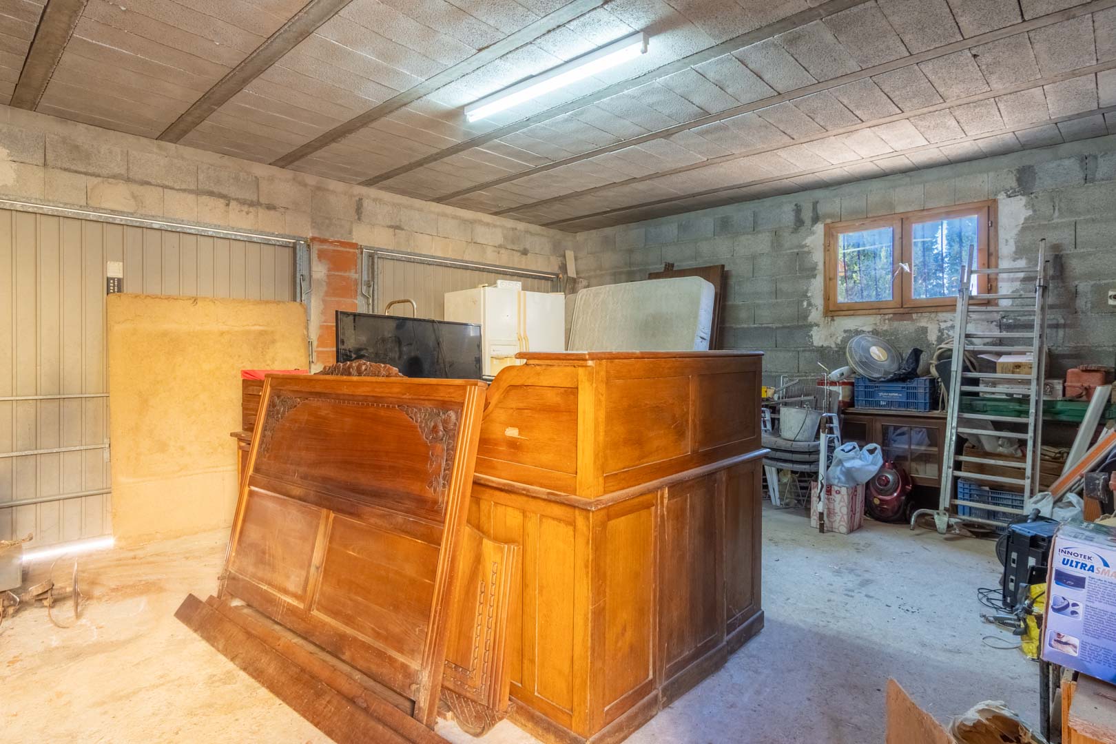 Grand espace de rangement ou garage avec murs en parpaings, plafond à poutrelles, éclairage fluorescent et une fenêtre en bois apportant de la lumière naturelle.