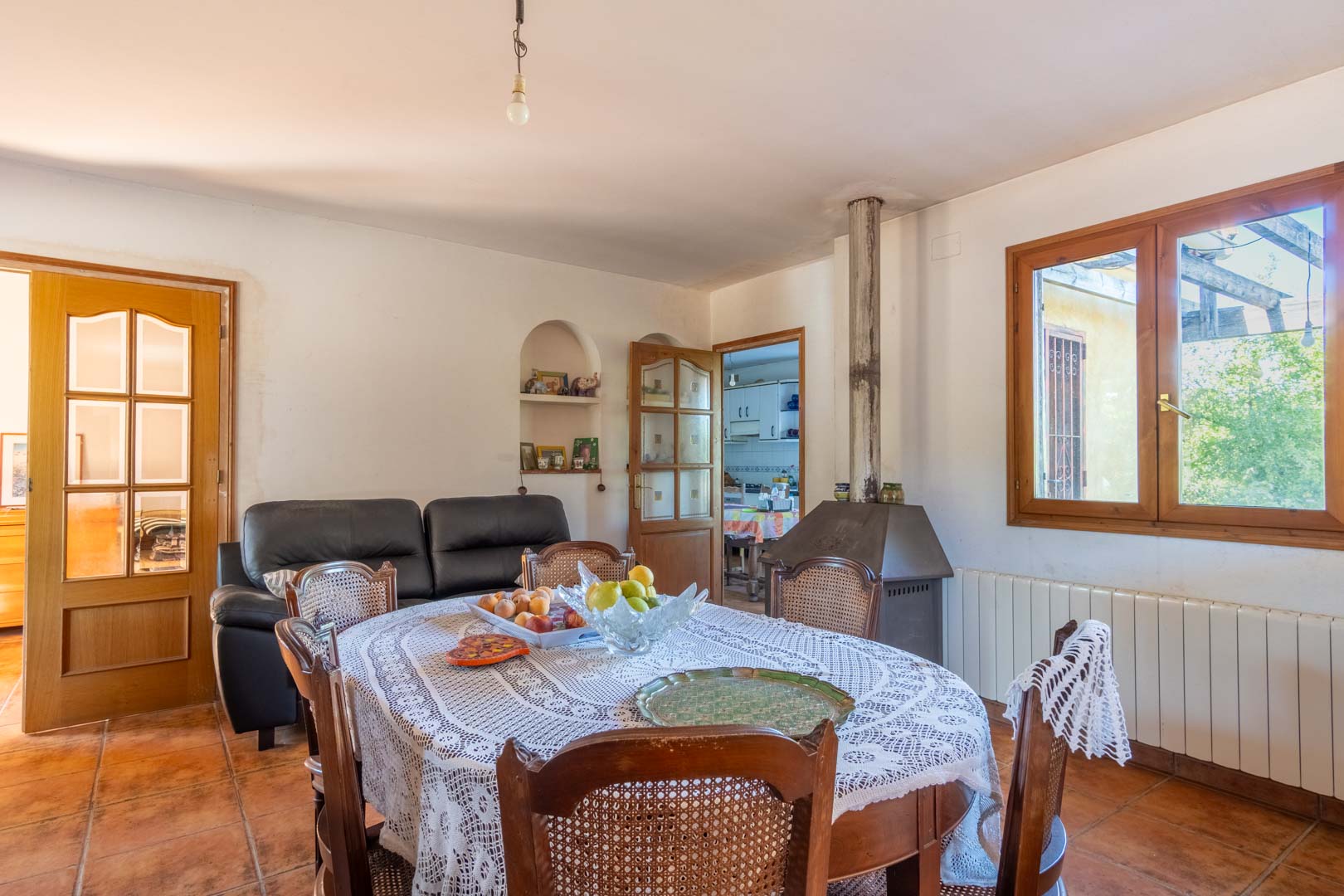 Salle à manger avec sol en terre cuite, poêle à bois métallique et mobilier en bois classique. La pièce lumineuse offre un accès direct à la cuisine et dispose d'un radiateur.