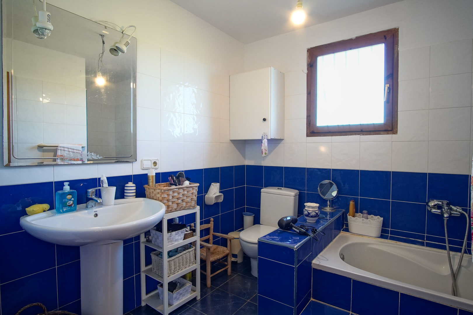 Salle de bain aux carreaux bleus et blancs, comprenant un lavabo sur colonne, une baignoire encastrée et une fenêtre à cadre en bois.