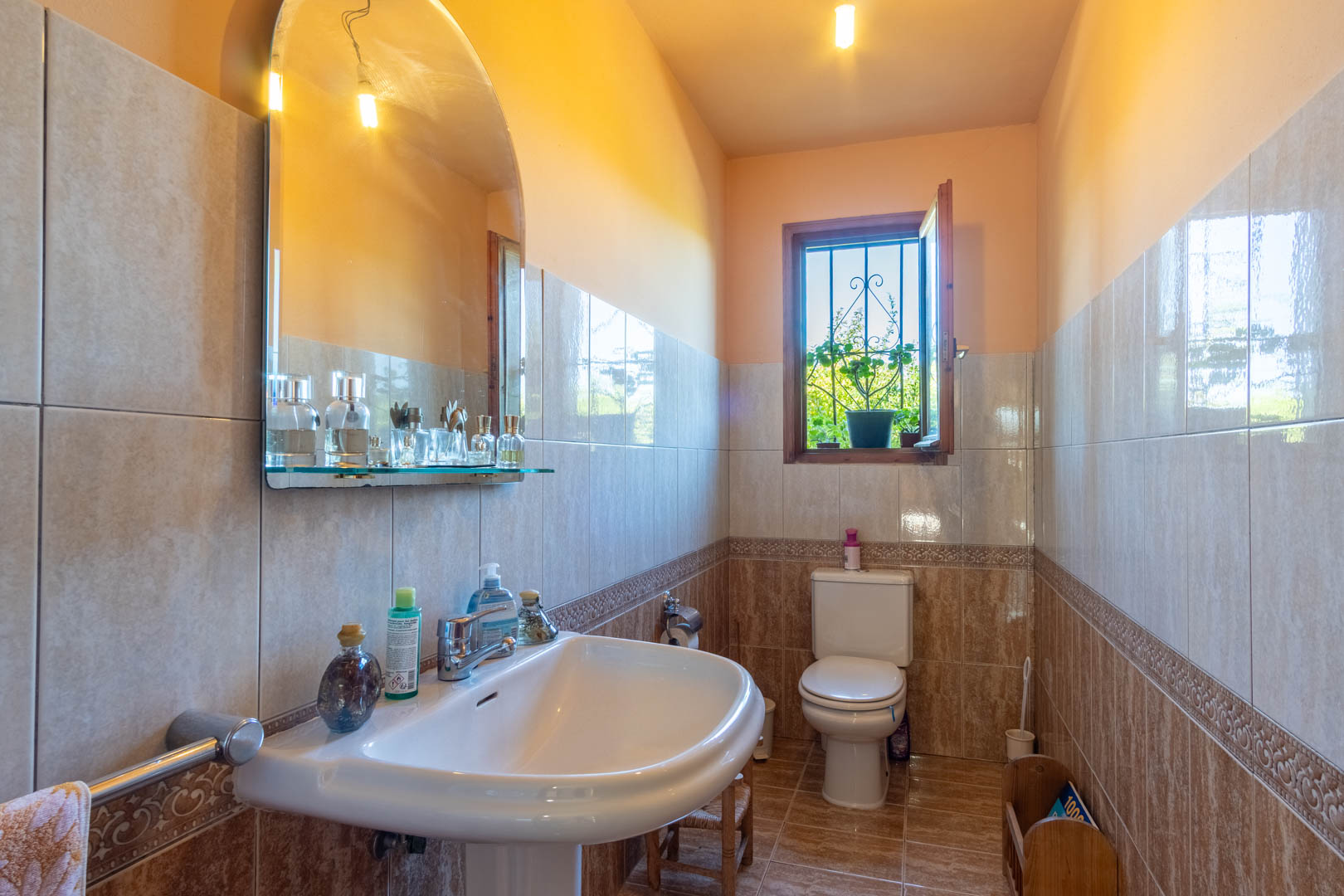 Salle de bain avec carrelage mural aux tons terre, lavabo sur colonne, miroir cintré et fenêtre en bois offrant une lumière naturelle.