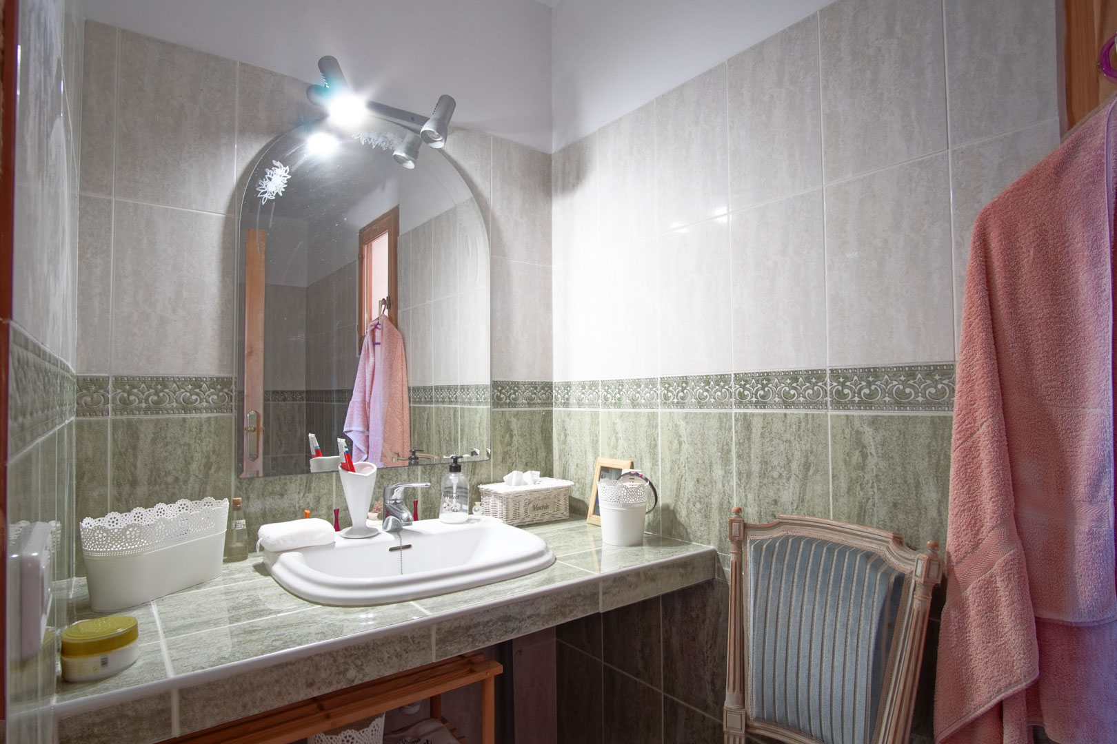 Salle de bain avec plan de travail carrelé vert, lavabo encastré, miroir cintré avec spots intégrés et murs ornés d'une frise décorative.