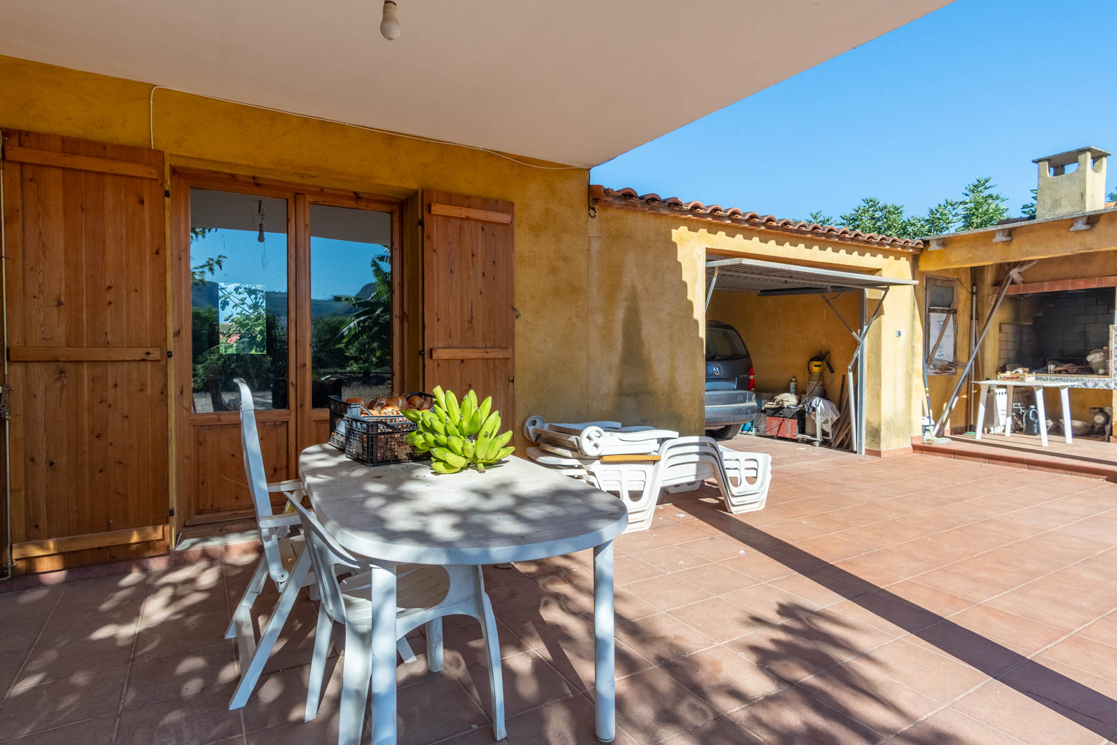 Terrasse extérieure avec carrelage en terre cuite, espace repas ombragé et accès au garage couvert ainsi qu'à la cuisine d'été avec barbecue.