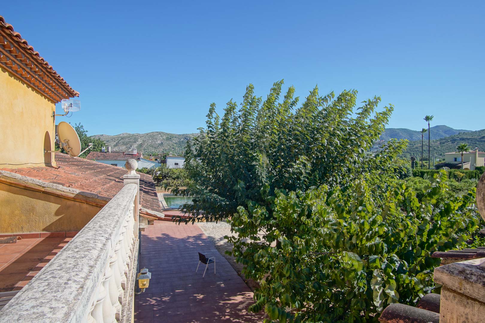 Vue depuis la terrasse supérieure avec balustrade en pierre, dominant le patio, la piscine et la végétation luxuriante avec les montagnes en arrière-plan.
