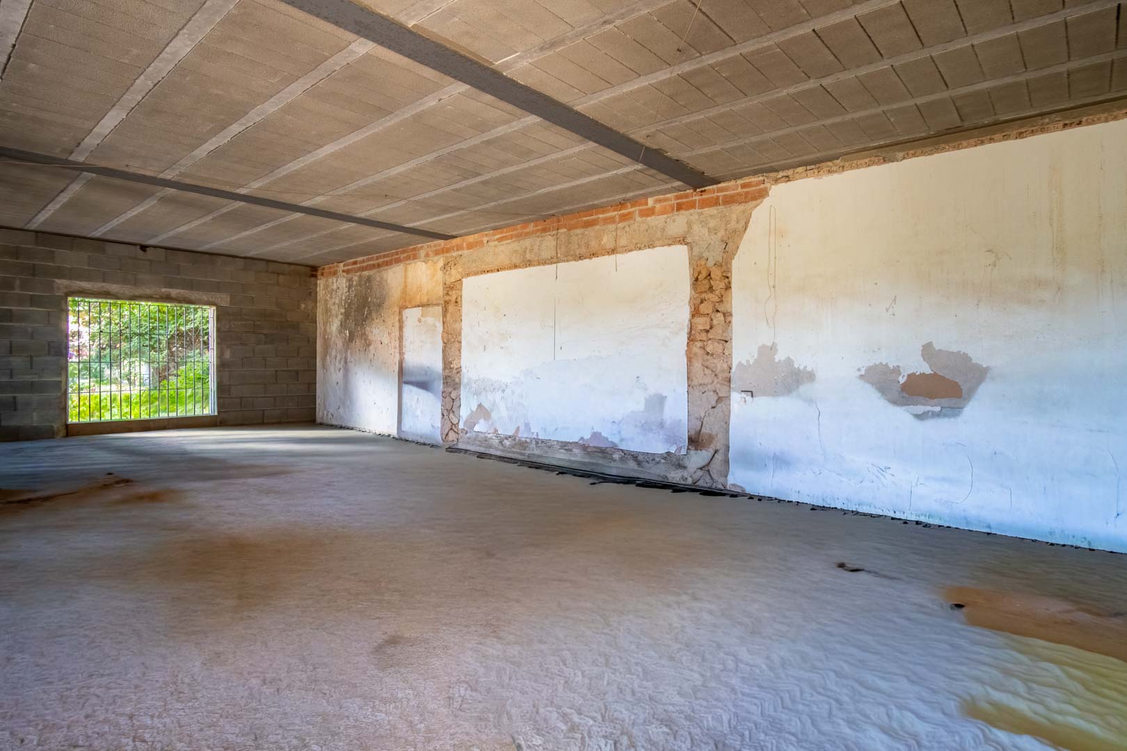 Espace intérieur ouvert en cours de rénovation avec sol en béton, poutres apparentes au plafond et murs mêlant pierre d'origine et briques.