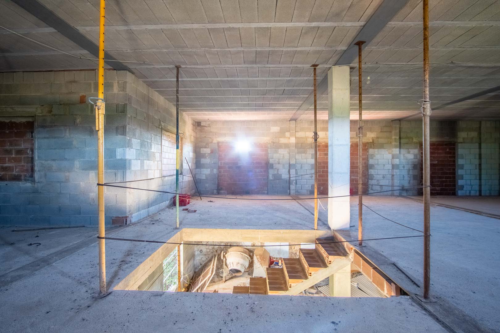 Intérieur ouvert en cours de construction avec murs en parpaings, poutres métalliques et trémie d'escalier communiquant avec le niveau inférieur.