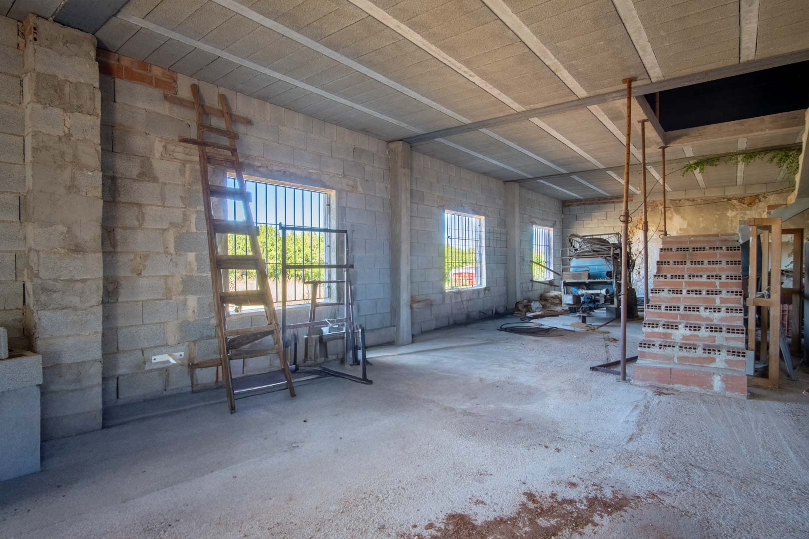 Intérieur spacieux et ouvert en cours de rénovation, avec des murs en parpaings, un plafond à poutrelles et des fenêtres grillagées laissant entrer la lumière.