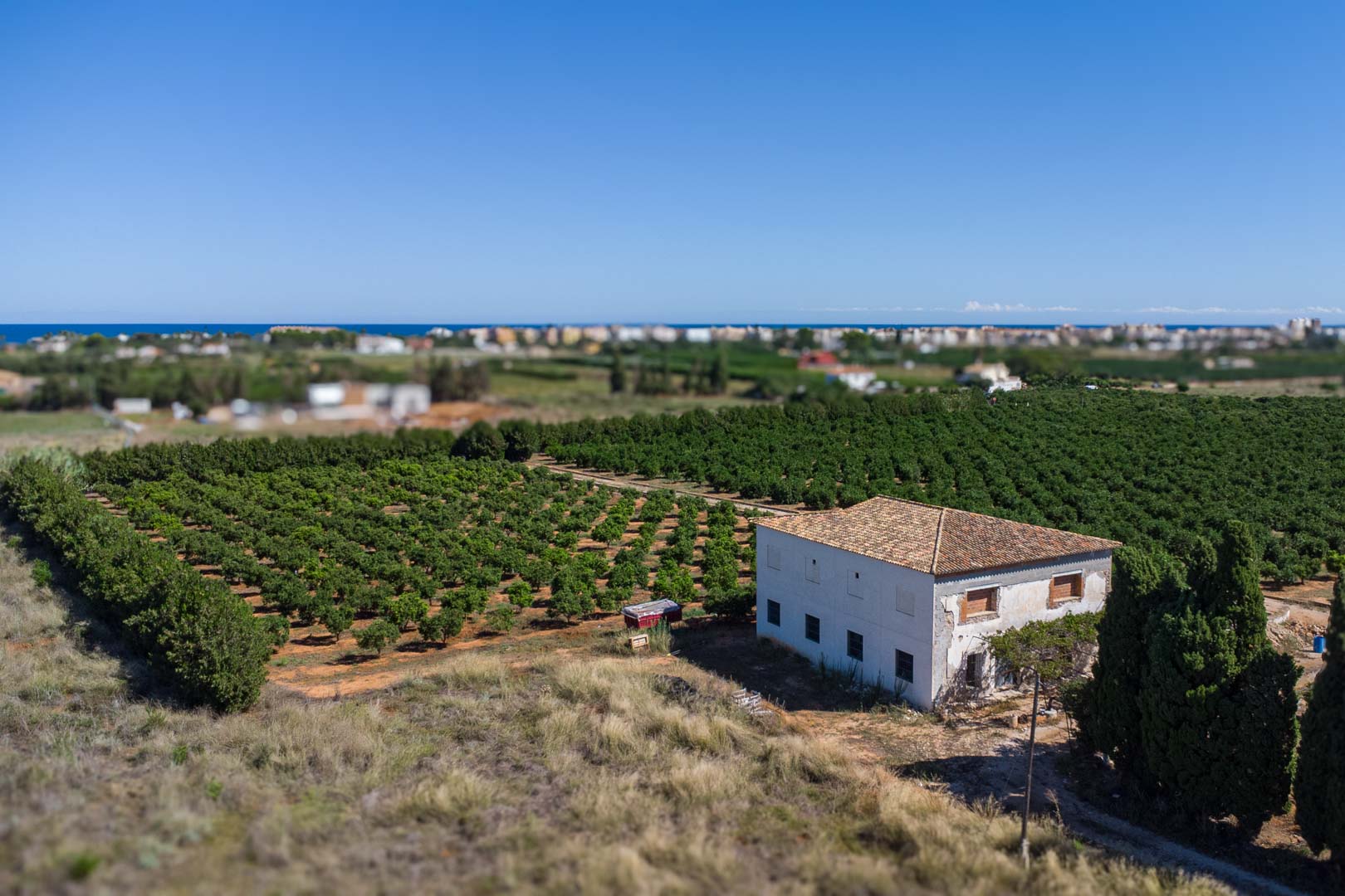 Vue aérienne de la maison de campagne entourée de plantations, avec sa façade blanche, son toit en tuiles et une vue dégagée sur la mer Méditerranée.