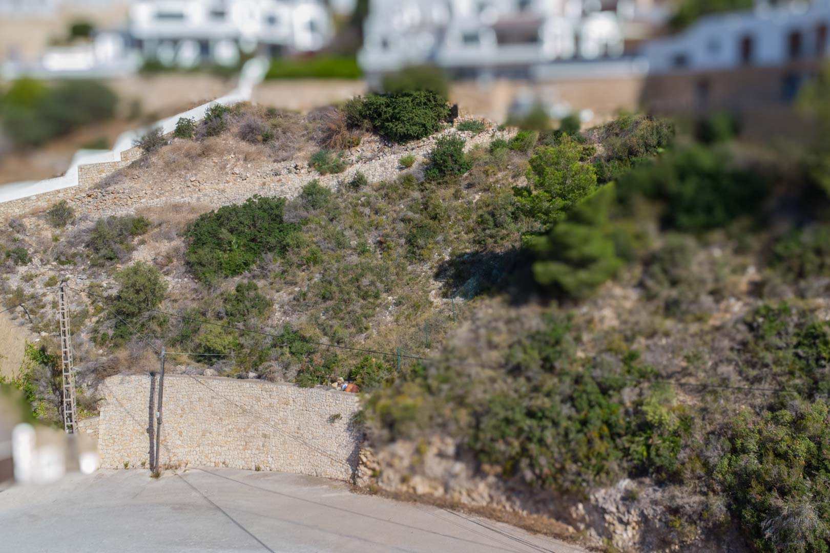 Terrain urbain surélevé à El Portet avec des murs de soutènement en pierre naturelle et une végétation méditerranéenne bordant une route goudronnée.