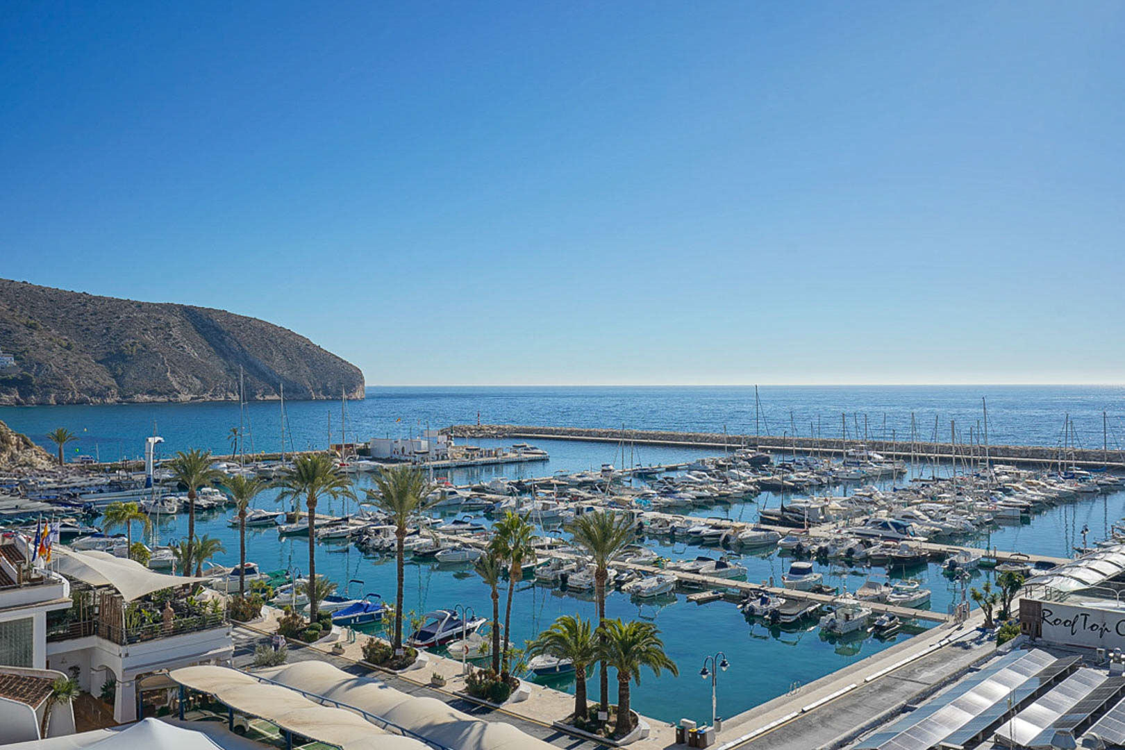 Vue panoramique sur le port de plaisance de Moraira avec ses nombreux bateaux, ses palmiers et la falaise côtière en arrière-plan.