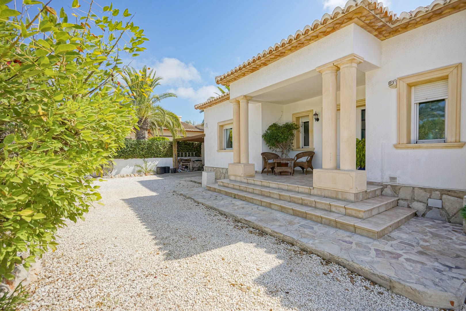 Entrée de la villa avec gravier blanc, porche à colonnes et mobilier en osier.