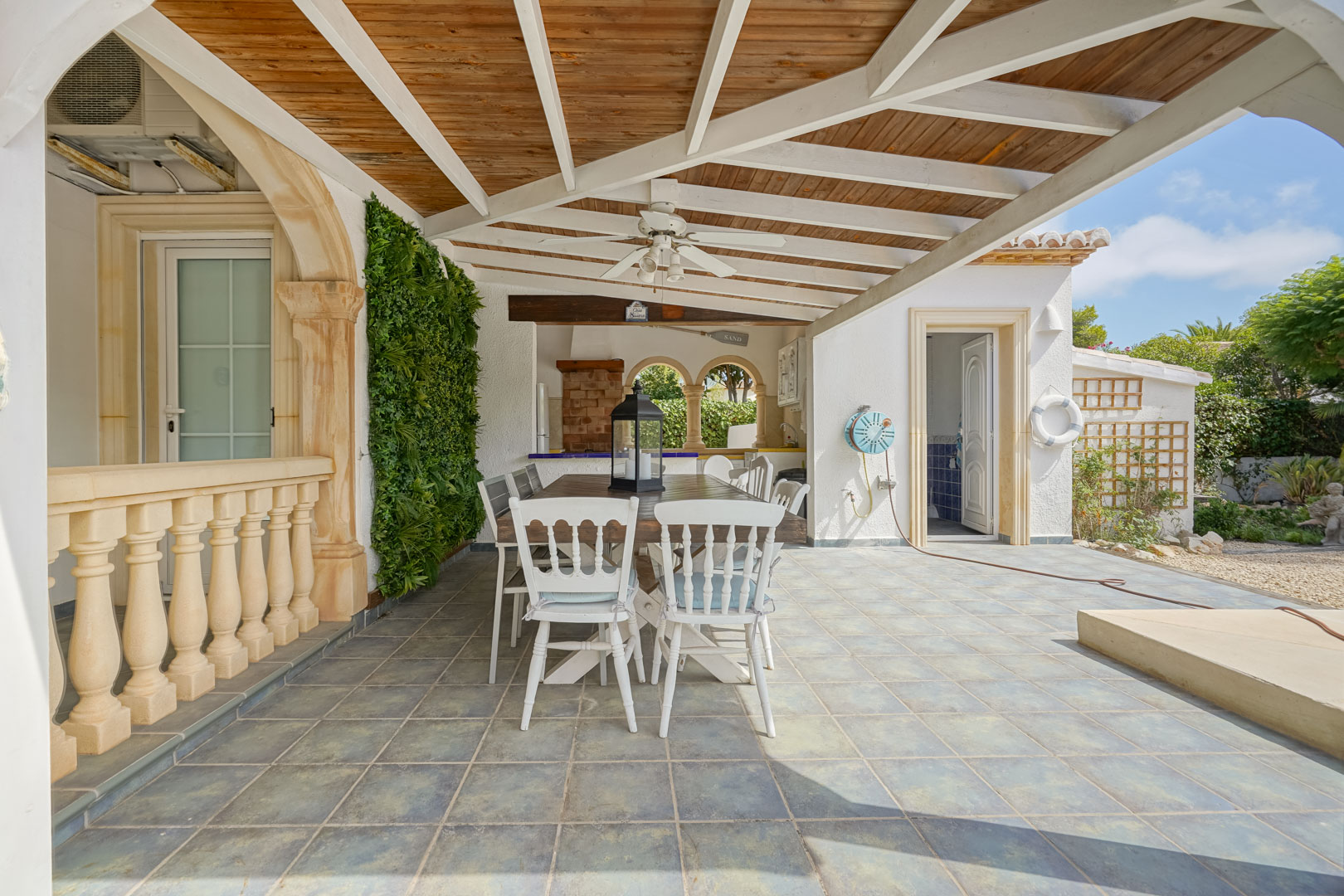 Salle à manger extérieure couverte avec table en bois et chaises blanches, à côté d'une cuisine extérieure avec barbecue.