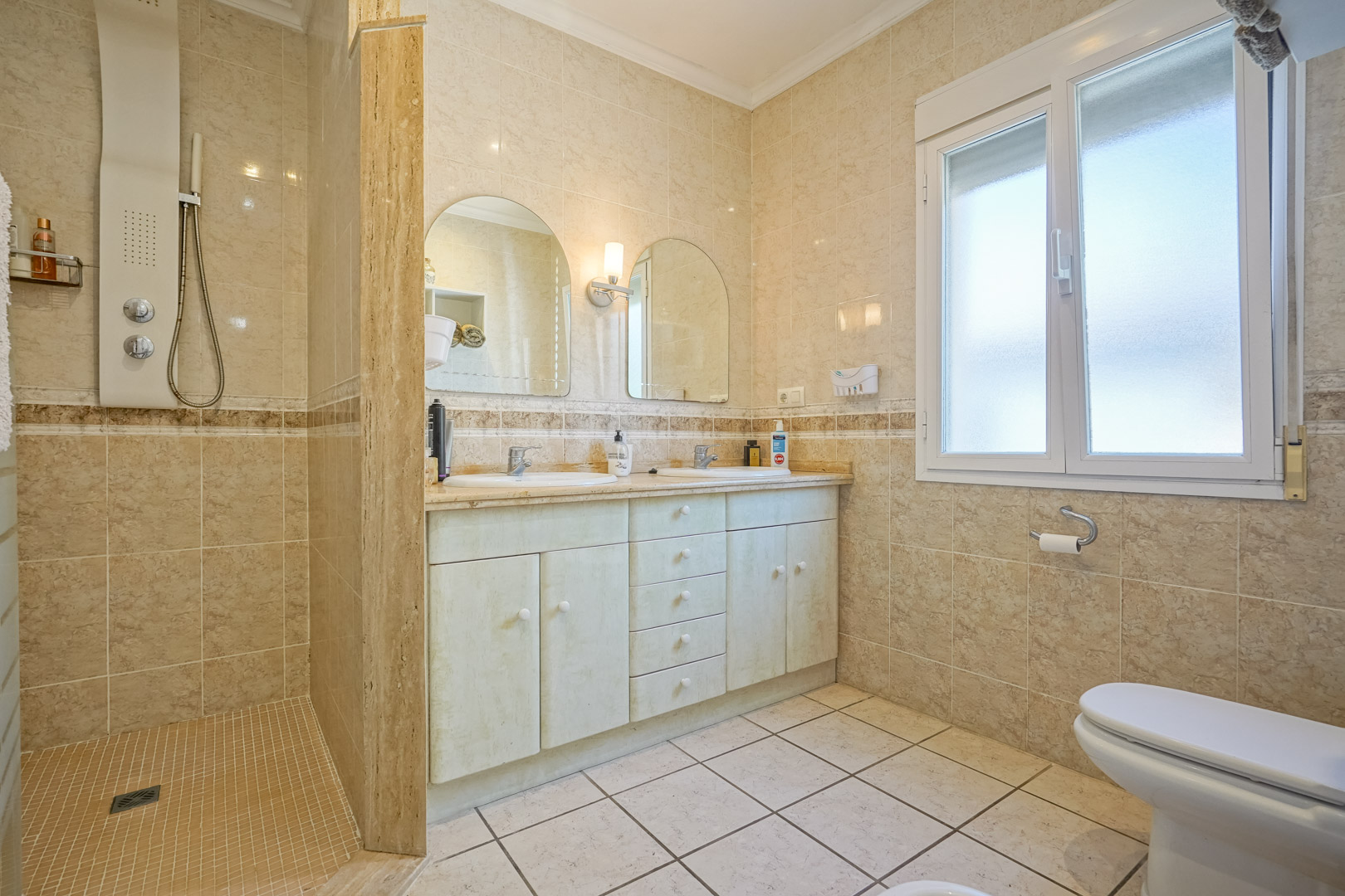 Salle de bain avec douche à l'italienne, meuble-lavabo double vasque et carrelage crème.