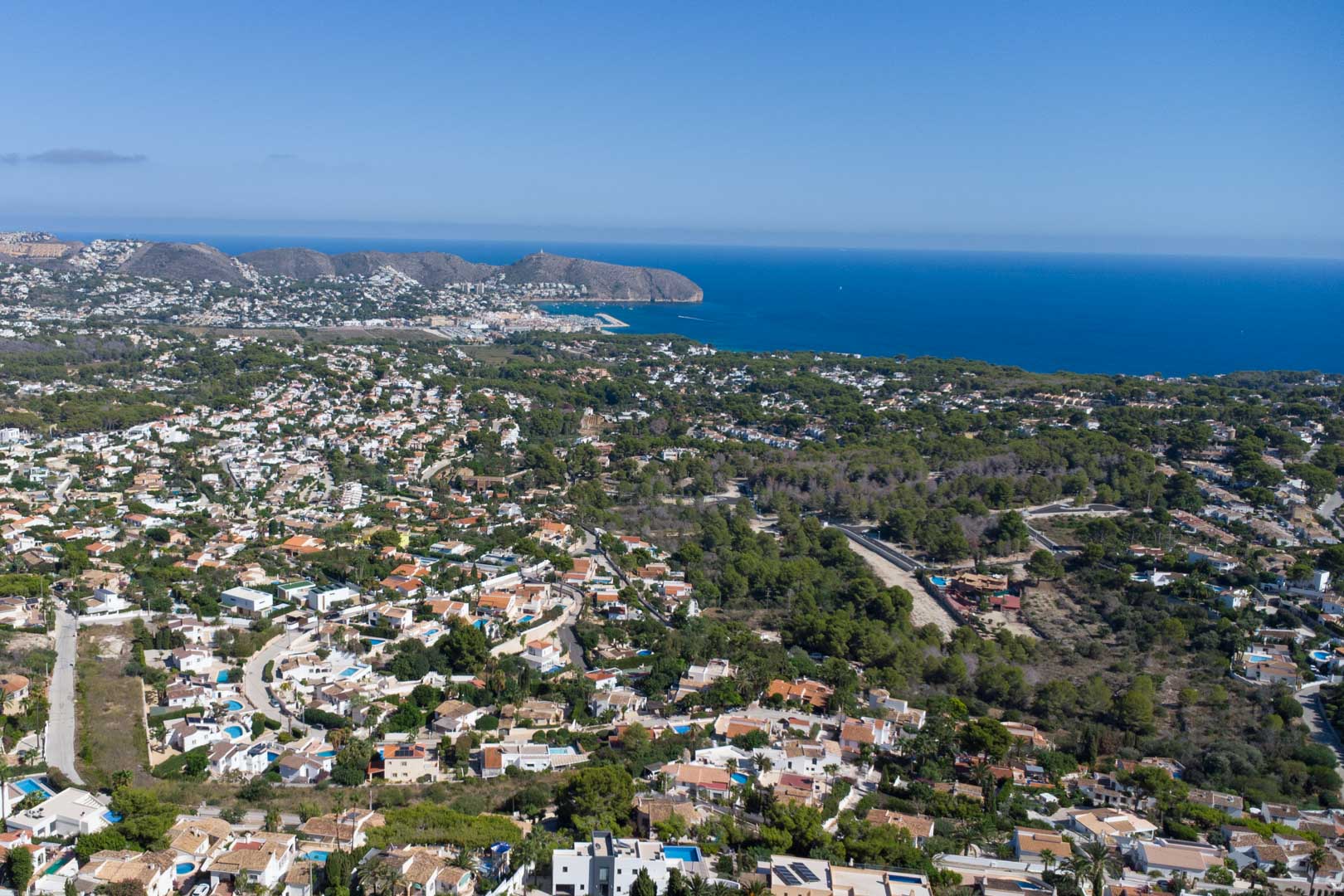 Vue aérienne panoramique de Moraira révélant le littoral, le port et le Cap d'Or, entourés de quartiers résidentiels et de forêts de pins méditerranéens.