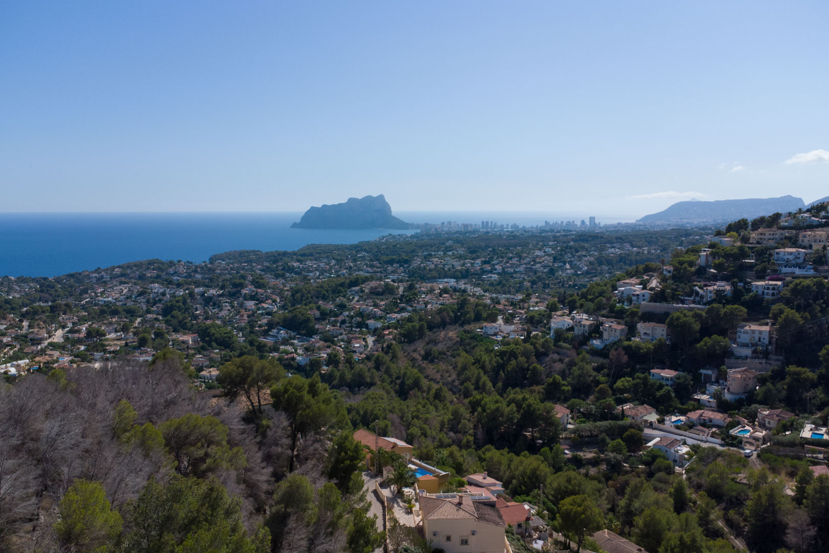 Vue panoramique surélevée sur la mer Méditerranée et le Peñón de Ifach, illustrant le paysage côtier verdoyant et les collines résidentielles.