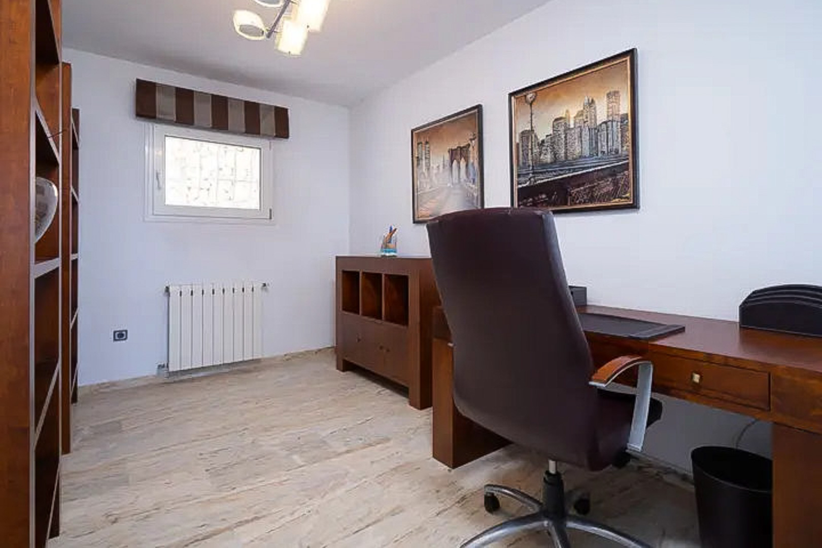 Bureau lumineux avec mobilier en bois, bureau fonctionnel et chaise en cuir. Comprend des étagères, un sol en pierre claire et des tableaux de paysages urbains.
