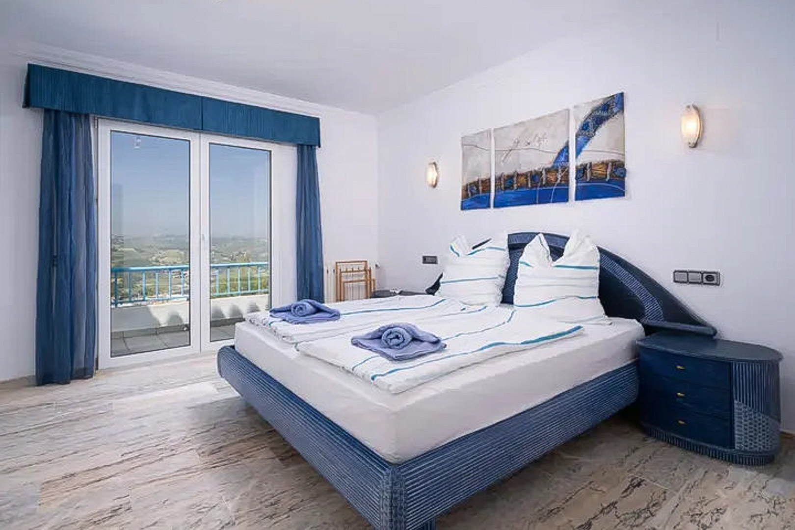 Chambre avec sol en marbre, lit double aux tons bleus et accès direct à un balcon offrant une vue dégagée sur le paysage méditerranéen.