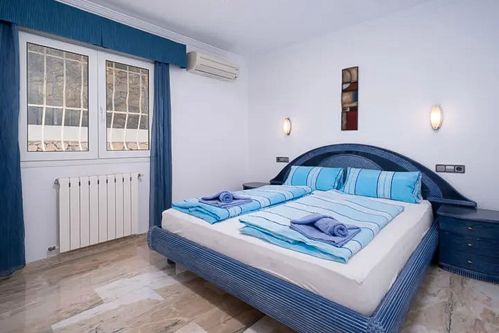 Chambre avec sol en marbre poli, lit double aux tons bleus, climatisation murale, radiateur et fenêtre sécurisée avec rideaux assortis.