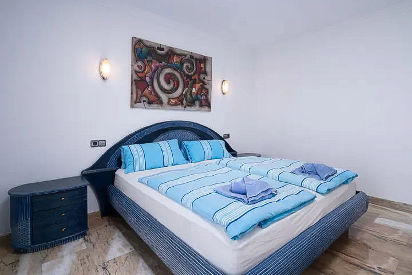 Chambre avec tête de lit en osier bleu et table de chevet assortie. Sol en marbre poli, appliques murales et tableau abstrait au-dessus du lit.