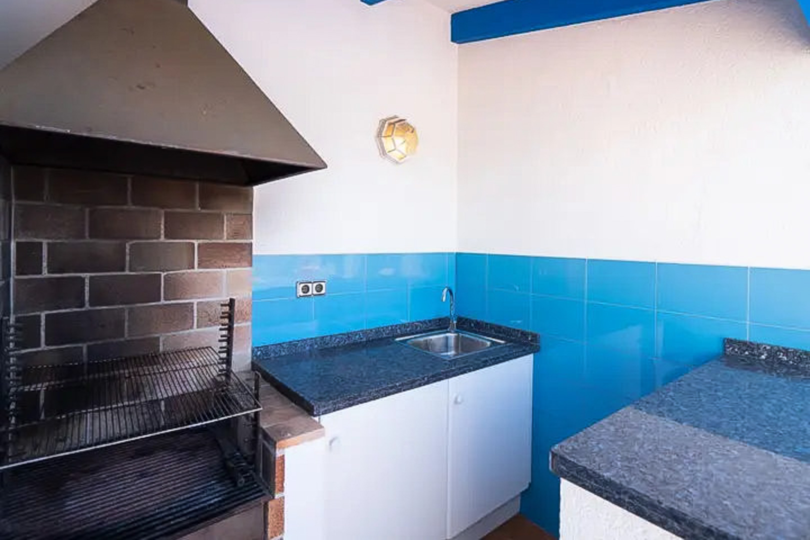 Espace barbecue extérieur avec hotte métallique, plan de travail en granit sombre, évier en inox et crédence en carreaux bleu méditerranéen.