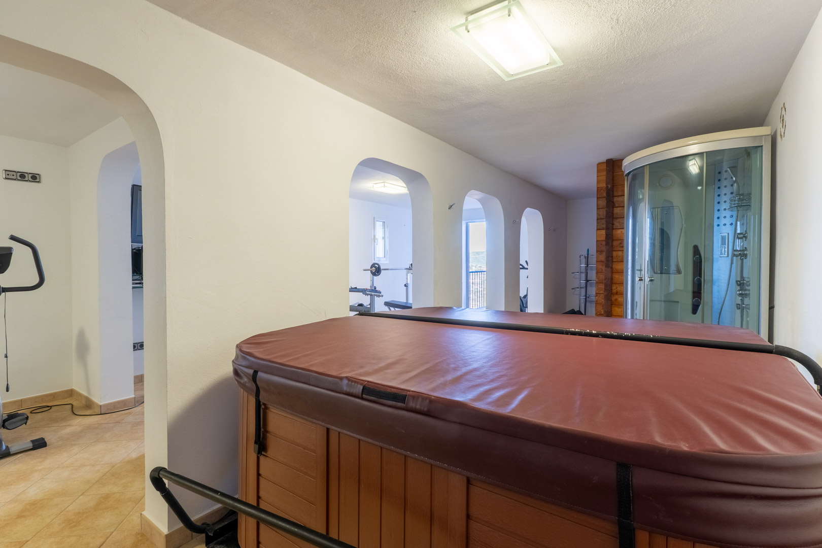 Espace bien-être intérieur comprenant un jacuzzi avec couverture, une cabine de douche d'hydromassage et des ouvertures en arche vers la salle de sport.