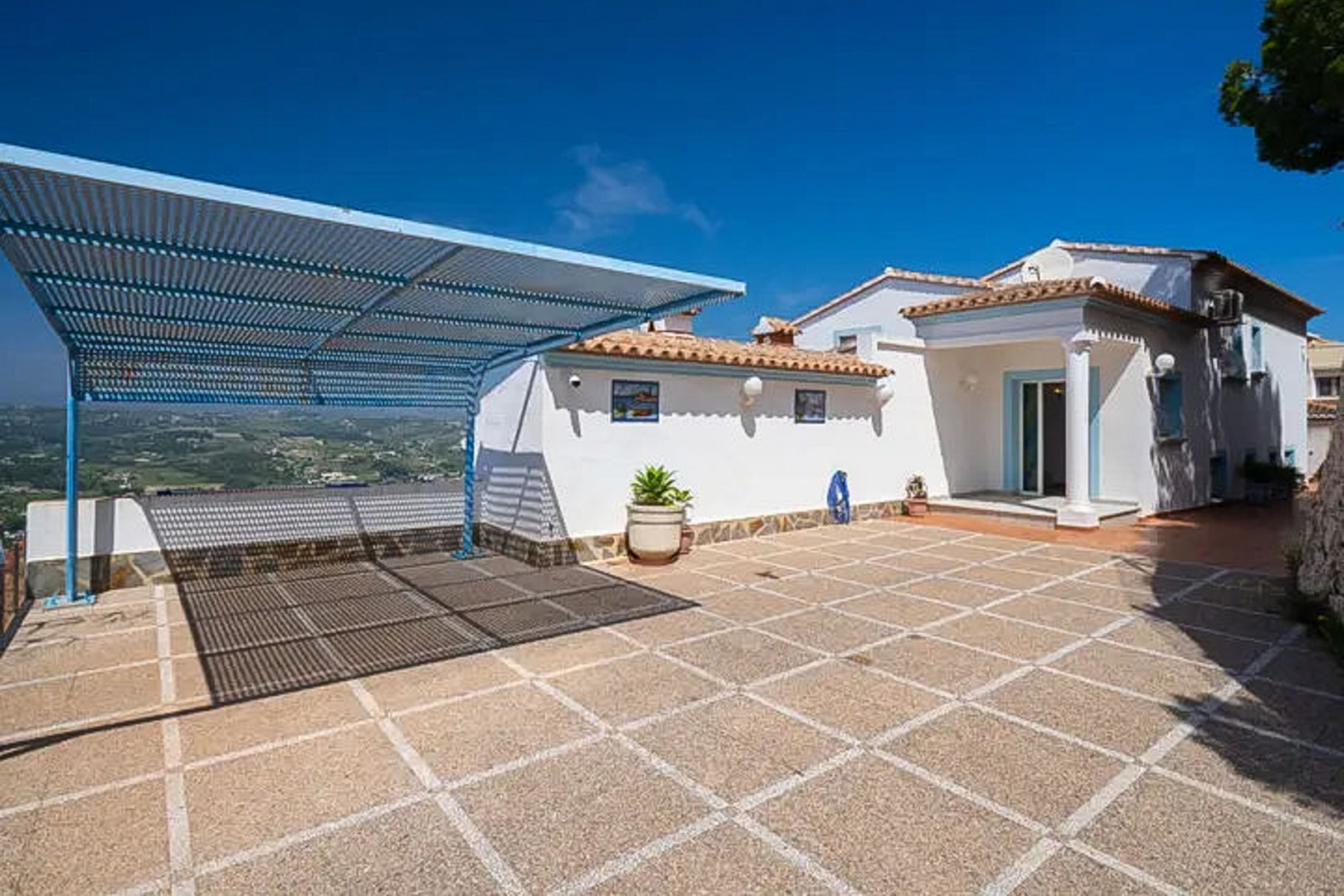Espace de stationnement extérieur avec revêtement carrelé, abri de voiture métallique bleu et vue dégagée sur les montagnes environnantes de la Costa Blanca.