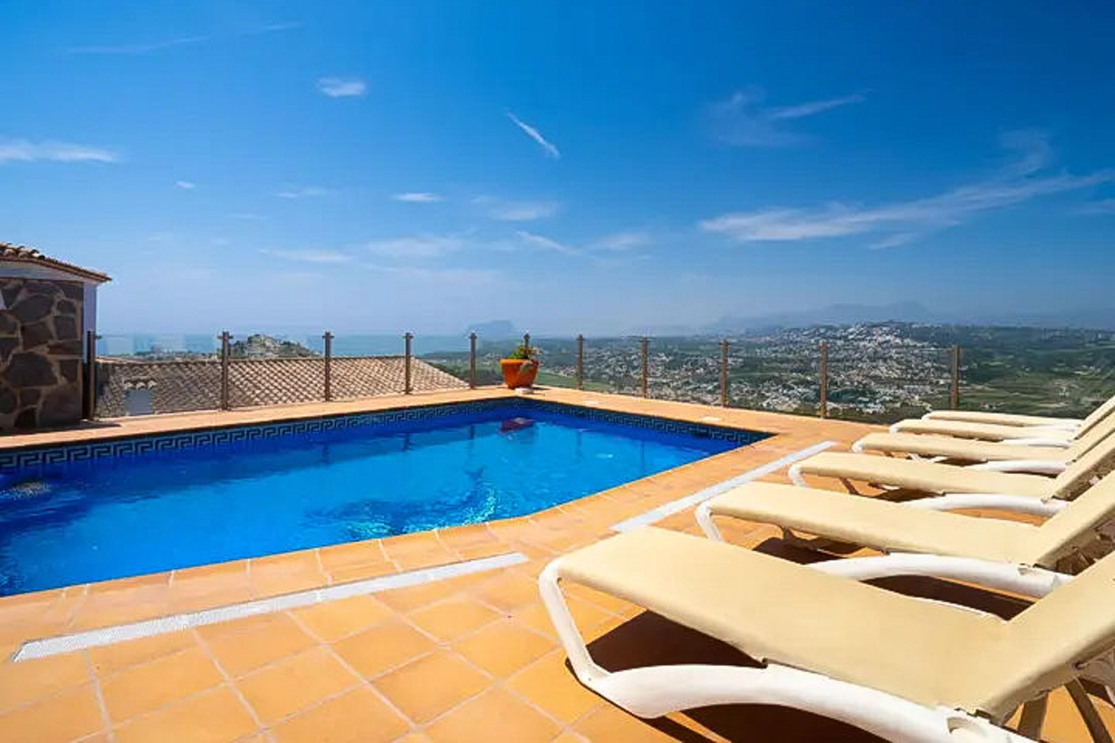 Piscine extérieure avec frise grecque, terrasse en terre cuite équipée de transats et garde-corps en verre offrant une vue panoramique sur la mer et Moraira.