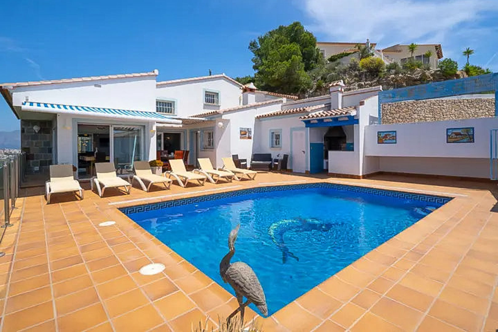 Piscine extérieure avec mosaïque de dauphin et terrasse en terre cuite, bordée de bains de soleil et d'une cuisine d'été intégrée à la villa.