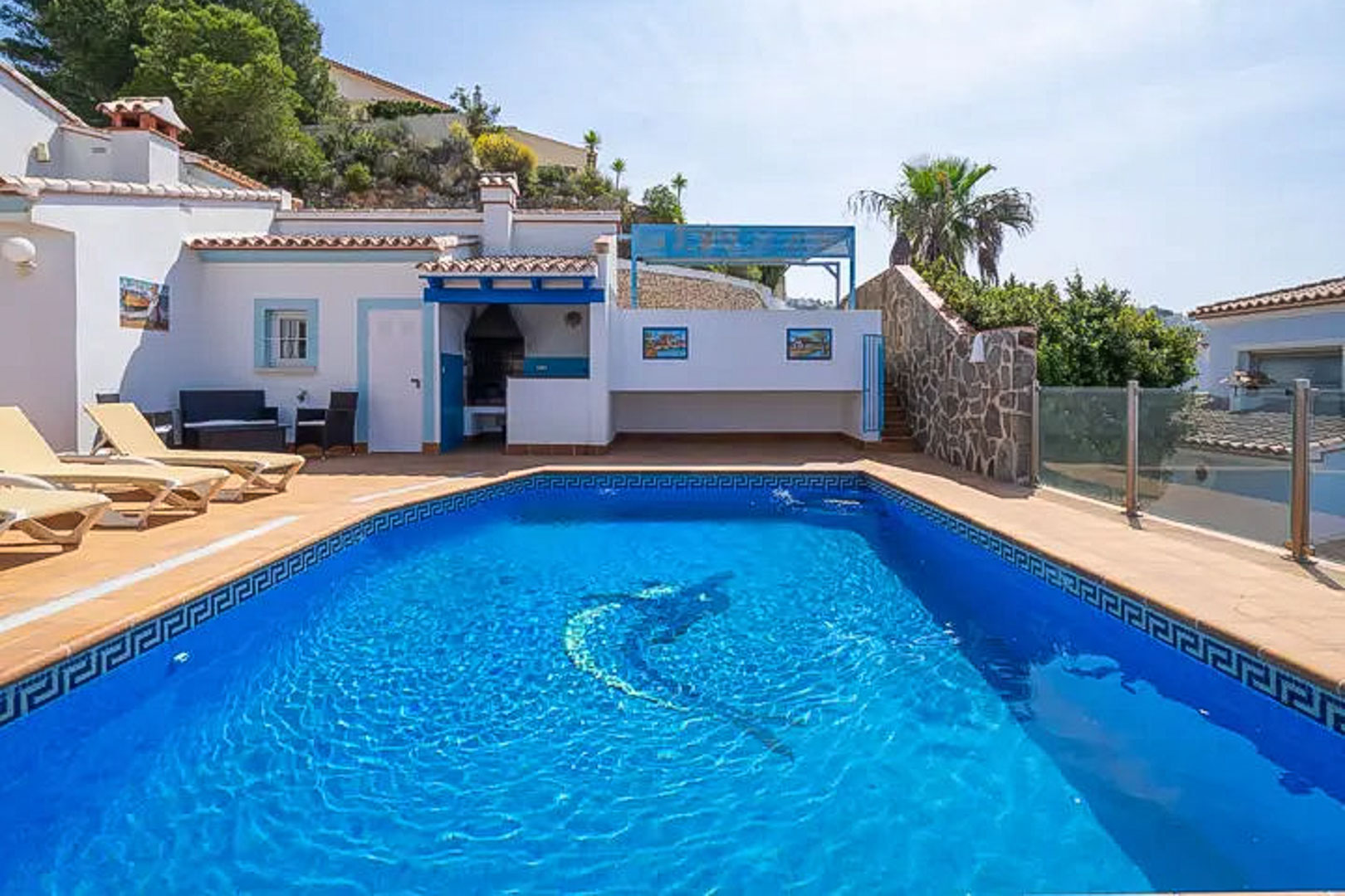 Piscine extérieure avec mosaïque de dauphin, terrasse en terre cuite avec bains de soleil et un espace barbecue couvert aux finitions bleues typiques.